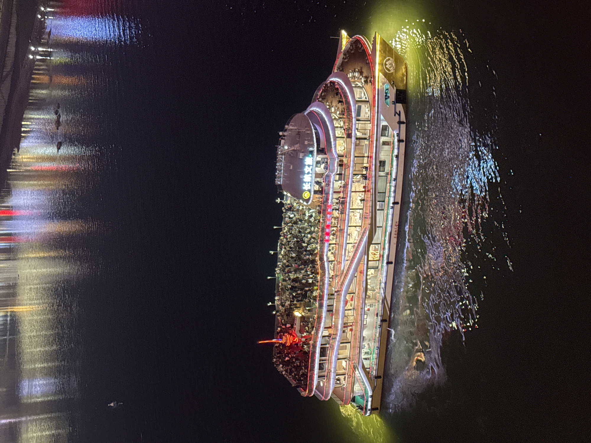 At night, a multi-story tour boat illuminated with decorative lights is sailing on a river near Yuzhong District, China. The boat is packed with people on its decks. Reflections of other lights are visible in the dark water on the opposite bank. Ripples and churning water are near the boat, suggesting it is moving. The immediate area around the boat is illuminated by green and gold lights. The overall scene is dark.