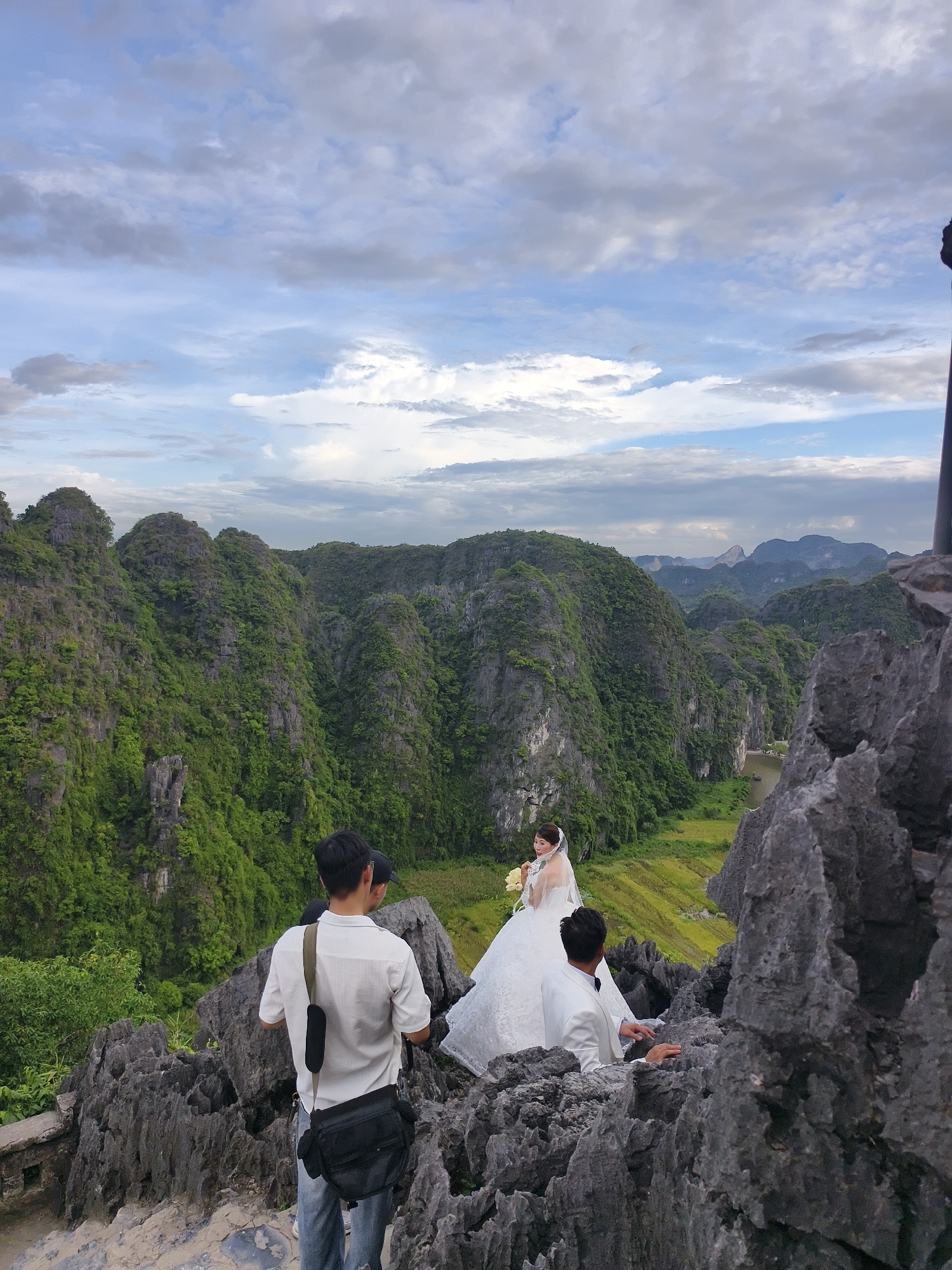The image depicts a wedding photoshoot taking place on a rocky outcrop overlooking a valley of karst mountains in Hoa Lư Ward, Vietnam. In the foreground, three individuals are visible. One person, a male, is wearing a white short-sleeved shirt and blue jeans and is carrying a black shoulder bag. He is standing on the rocky terrain, looking towards the valley. Another person, also male, is wearing a white suit and appears to be interacting with the bride. The bride, positioned further into the scene, is wearing a white wedding dress and veil, holding a bouquet of yellow flowers. She is looking towards the right of the frame. The background is dominated by lush green, jungle-covered karst mountains stretching into the distance, with a body of water visible in the valley floor. The sky is partly cloudy with a mix of blue sky and white and grey clouds. The overall scene captures a romantic wedding moment set against a dramatic natural landscape.