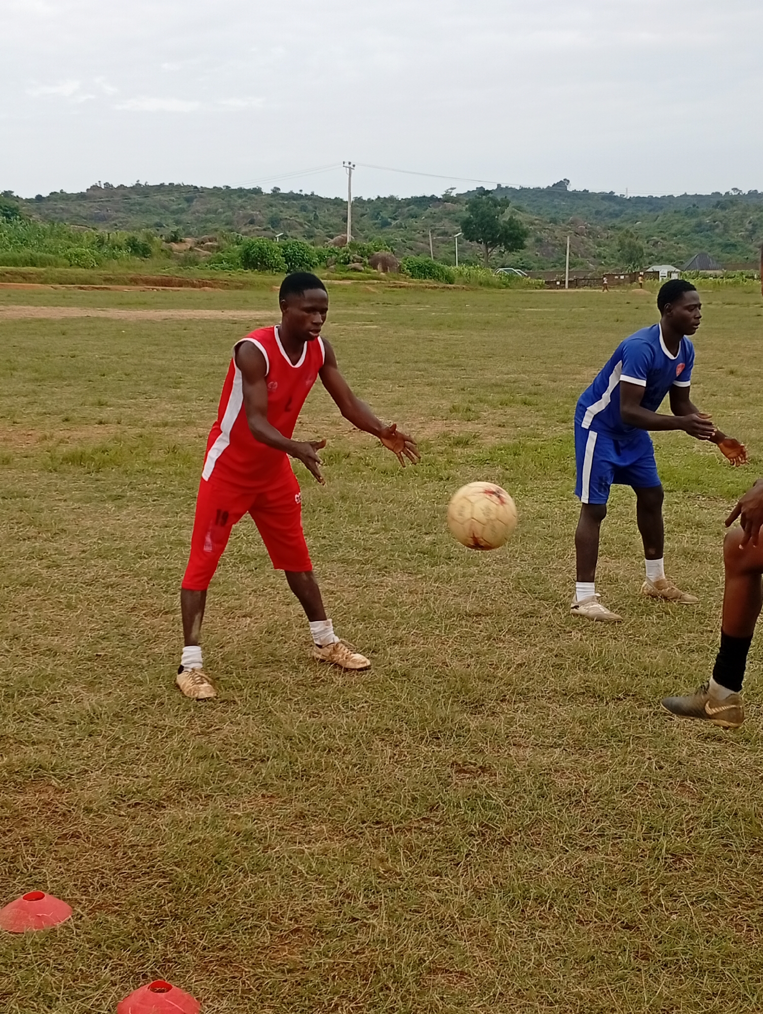 An outdoor daytime shot shows three young Black males engaged in a soccer training drill on a grassy field in Jos, Nigeria. The foreground features a young male in a red athletic uniform, including shorts and a sleeveless top with white trim. His arms are outstretched as if preparing to receive or pass a ball. The middle ground is occupied by a second young male in a blue soccer uniform, including shorts and a t-shirt with white trim. He is also in a stance that suggests participation in the drill, with hands held in front of him. A scuffed, pale soccer ball is in mid-air between the two individuals, indicating an active moment in the drill. In the background, a third person, partially visible, is positioned to the right, with only their legs and feet visible in black socks and cleats. The environment consists of a vast, mostly flat grassy field with some dry patches. In the background, a hilly landscape with sparse green vegetation, scattered rocks, and a few scattered buildings can be observed. The sky is overcast with a light haze. The overall scene depicts a casual sports activity taking place in a rural or semi-rural area of Jos.