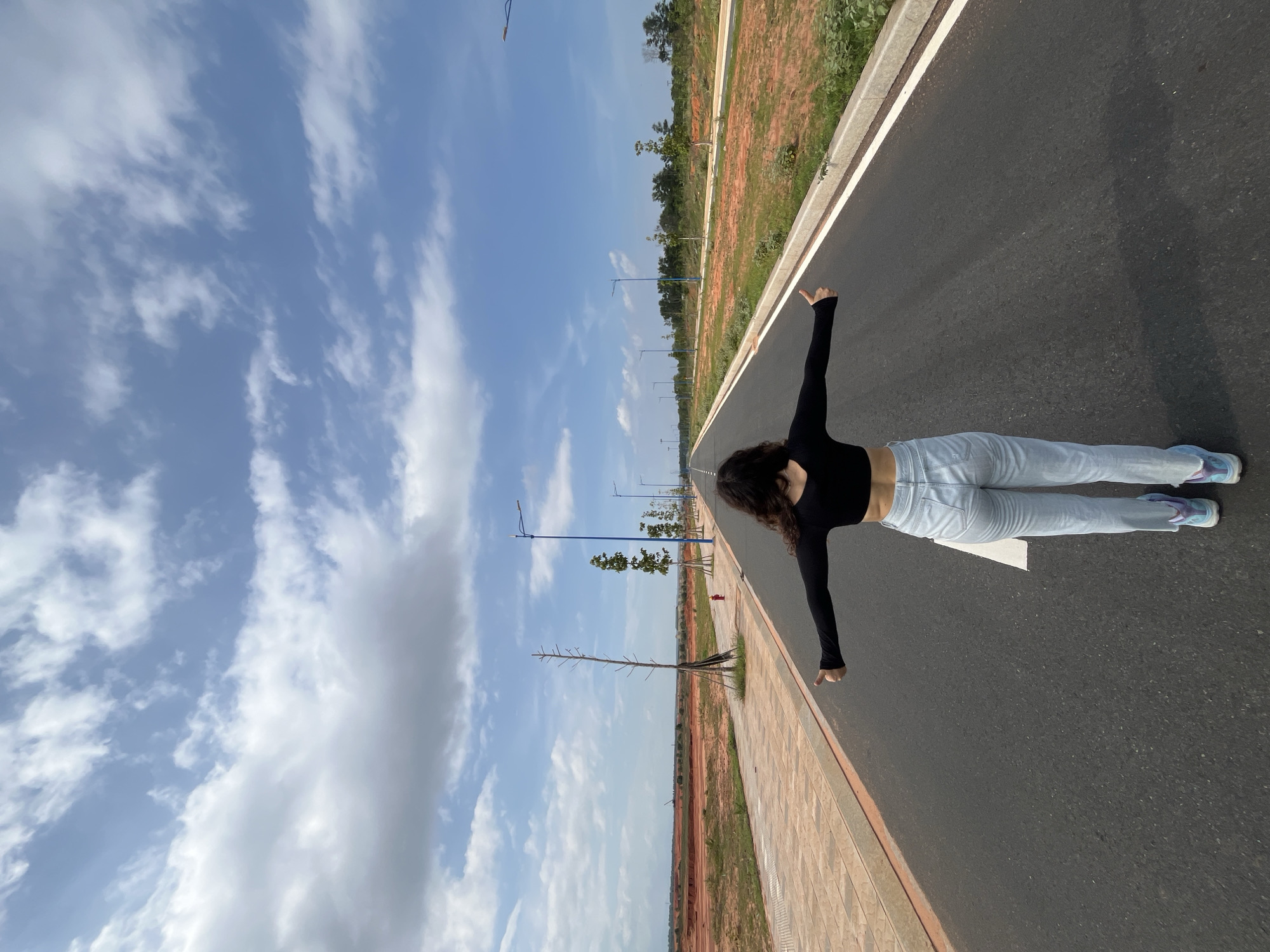 A person is standing on an asphalt road, facing away from the camera with their arms outstretched. They are wearing a black long-sleeve crop top and light-wash jeans. Their feet are clad in colorful sneakers. The road appears to be in a developing area, with construction sites and sparse vegetation visible on either side. There are streetlights lining the road at regular intervals. The sky is blue with scattered white clouds. The overall scene suggests an open, possibly rural or semi-urban landscape outside of Mui Ne, Vietnam.