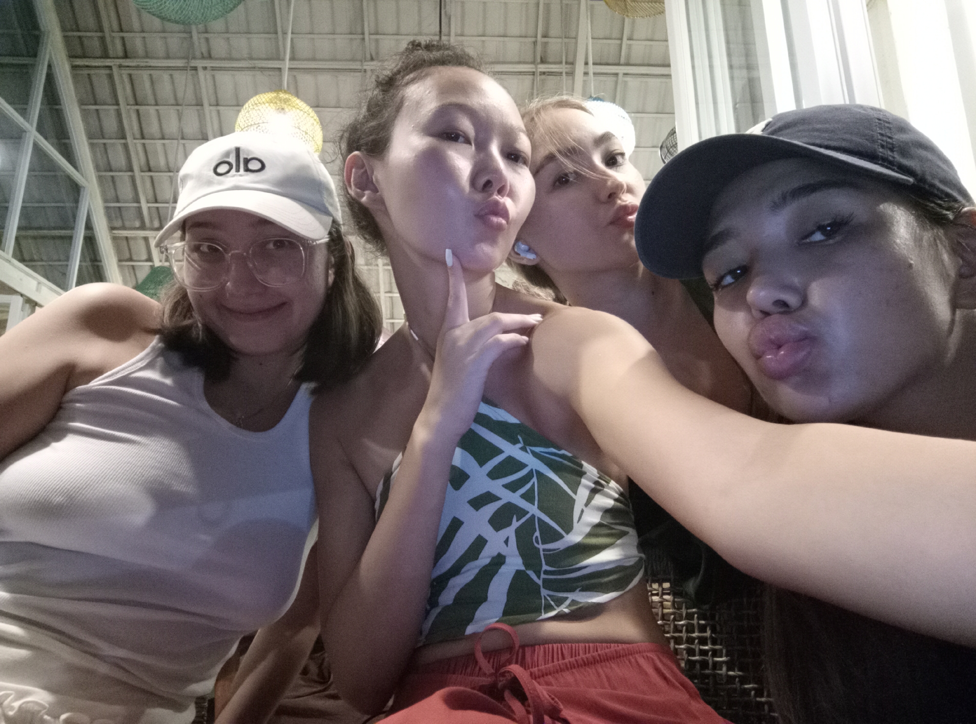 Four young women pose closely together for a selfie in what appears to be an indoor setting. The woman on the left wears a white tank top and a white baseball cap with "alo" printed on it, along with glasses. The woman in the center wears a halter top with a leaf pattern and red shorts. The third woman wears a white hair accessory and is positioned slightly behind the second woman. The woman on the right wears a dark baseball cap. All four women have pursed lips as if posing for a kiss. The location is identified as North Kuta, Indonesia. The environment appears to be a casual indoor space with a ceiling that has a grid pattern and woven decorative light fixtures.