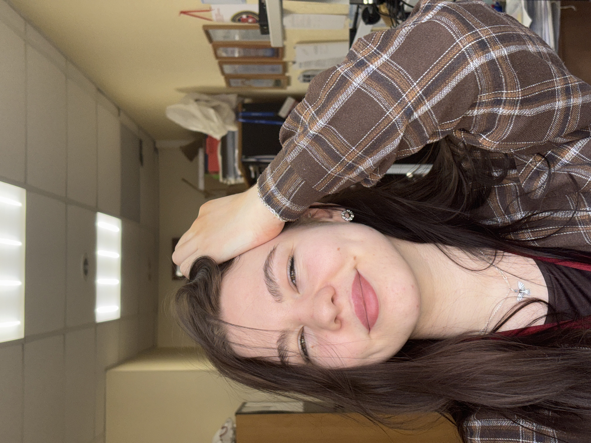 A young woman is indoors, wearing a brown plaid shirt and a silver necklace with a bird pendant. She is holding her head with her hand and smiling. The background includes an office desk with stacks of papers and office supplies, and a wall with fluorescent lighting. The location is presumed to be Perm, Russia, based on the prompt, but no specific details confirm this.
