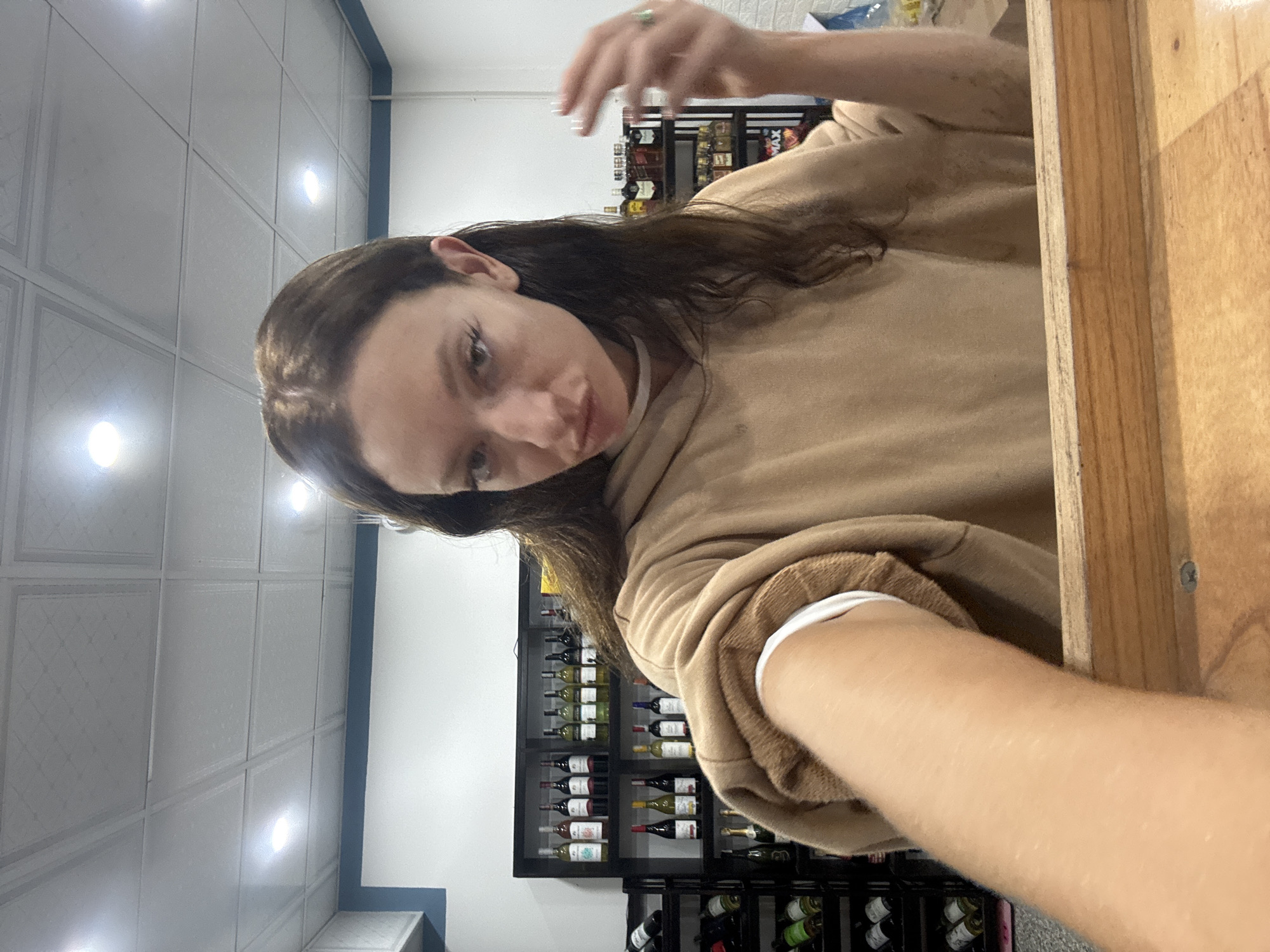 A woman with dark hair wearing a tan hoodie is in a store with wine racks on the wall. She is holding her hand up near her face, and part of a wooden shelf is visible to the right. The ceiling has recessed lighting. The location is likely in Đà Nẵng, Vietnam.