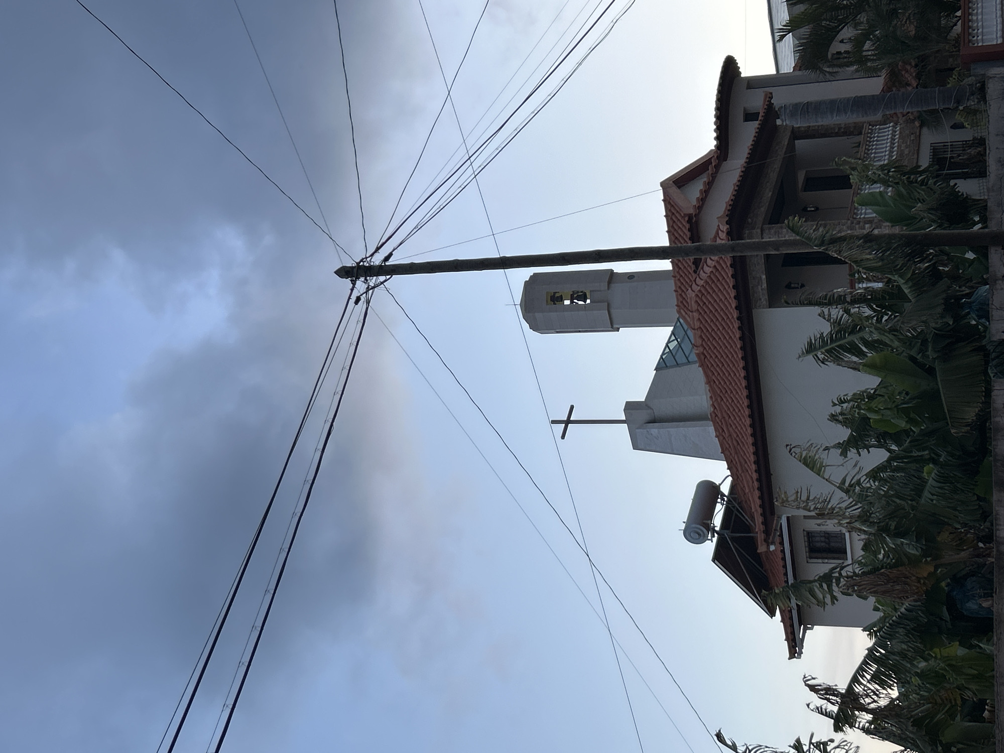 The image depicts a building with a cross and bell tower, likely a church, in Câmara de Lobos, Portugal. Power lines intersect the view. The building has a red tile roof and is partially obscured by green foliage. A water heater is mounted on the side. The sky transitions from cloudy to clear.