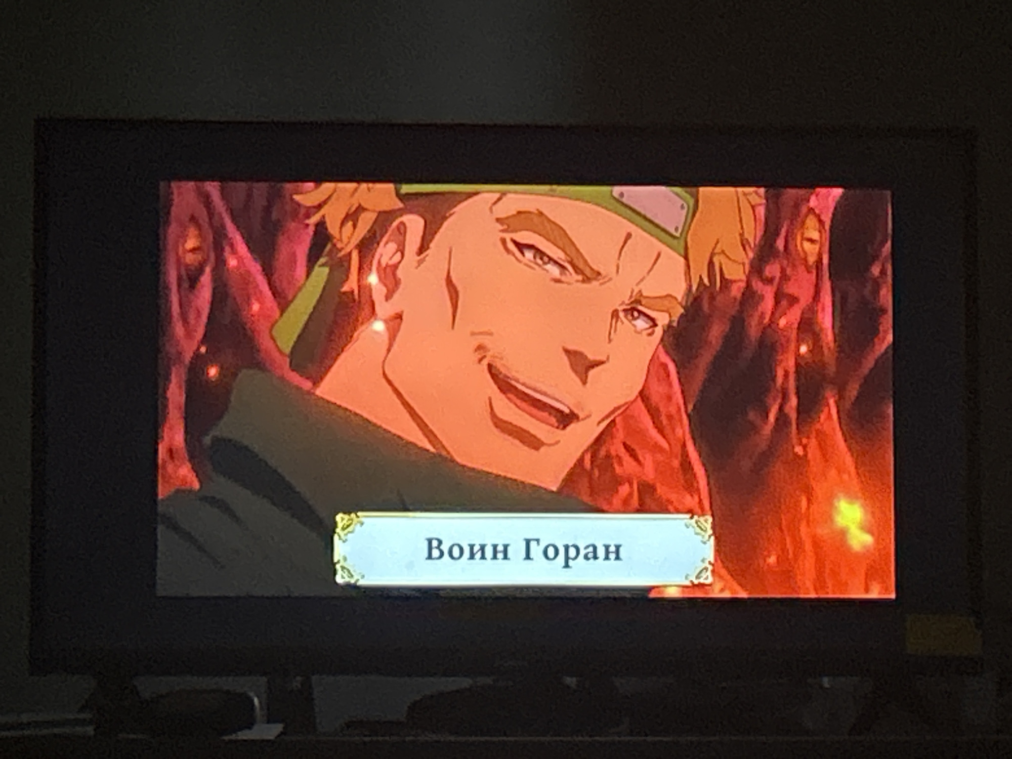 The image shows a close-up of a television screen displaying an anime-style character with orange hair and a green headband. The character has a smug expression and is looking to the right. Below the character is a light blue banner with decorative gold accents on the left and right. Inside the banner, in dark blue Cyrillic text, are the words "Воин Горан" which translates to "Warrior Goran". The background of the image on the screen is fiery red and orange, with indistinct shapes that suggest a dangerous or chaotic environment. The television screen is black and appears to be in a dimly lit room, with a subtle reflection visible on the screen's surface. There is no indication of Florești, Moldova in the image.