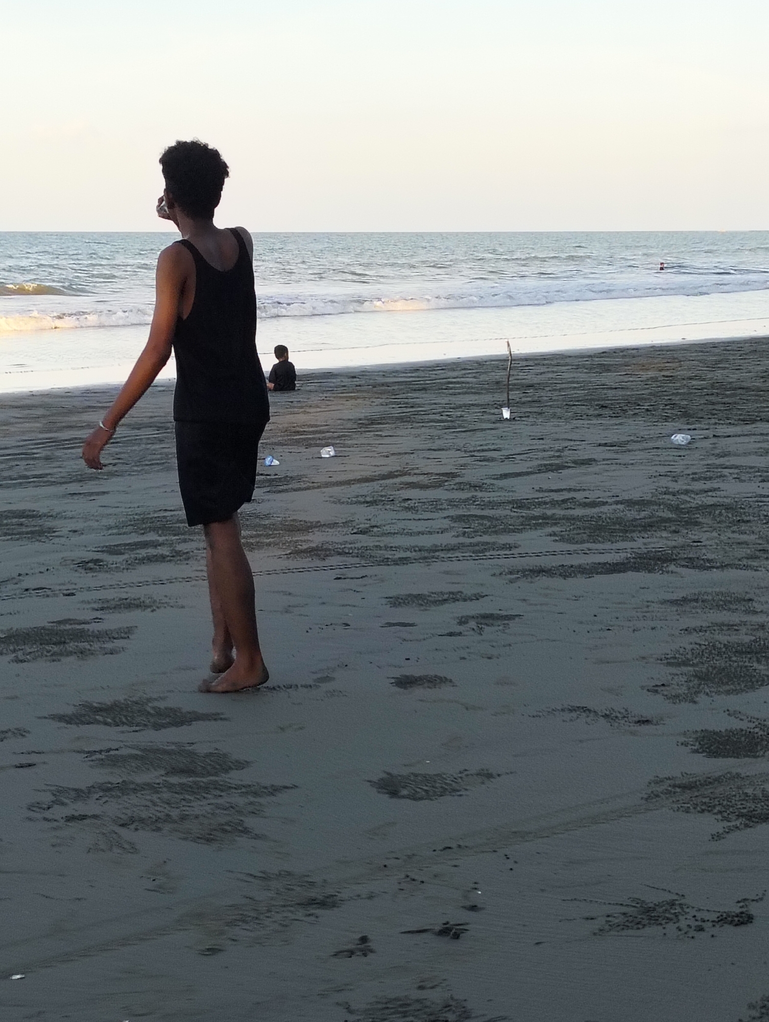 The image shows a beach scene at dusk, likely in Benteng, Indonesia, based on the presence of the sea and sandy shore. In the foreground, a young person, appearing to be male, stands with their back to the camera, facing the ocean. They are wearing a black tank top and black shorts and appear to be holding a phone or camera to their face, possibly taking a picture or recording. Their left arm is slightly outstretched. The person is barefoot and their feet are visible on the damp sand.
In the middle ground, a second, smaller figure, also appearing to be male, is seated further down the beach, facing away from the viewer and towards the ocean. They are wearing dark clothing.
The beach itself is sandy with some textured patterns in the sand, possibly from receding waves or footprints. There are a few scattered objects on the sand, including what appears to be a discarded plastic bottle and a thin stick with a white object attached near its base.
The ocean is visible in the background, with gentle waves breaking on the shore. The water appears calm, and the sky above is a pale, hazy blue, suggesting either early morning or late evening light. The horizon line where the ocean meets the sky is indistinct due to the haze. The overall atmosphere is peaceful and serene.