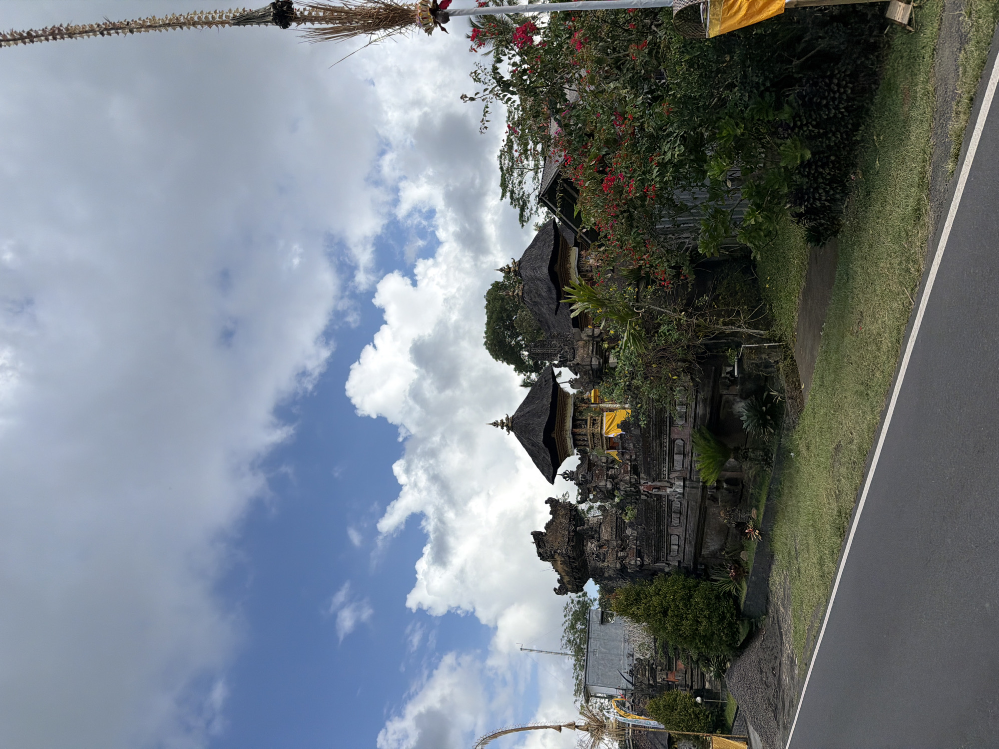 The image shows a traditional Balinese temple with intricate stone carvings and thatched roofs, located in Susut, Indonesia. The temple is situated along a road, with lush greenery and vibrant red flowers surrounding it. The sky is partly cloudy, with a mix of blue sky and white clouds. A tall, decorative bamboo pole with woven decorations stands on the right side of the frame. The road in the foreground is paved and has a white dividing line. The scene depicts a typical rural landscape in Bali, showcasing the island's rich cultural heritage and natural beauty.
