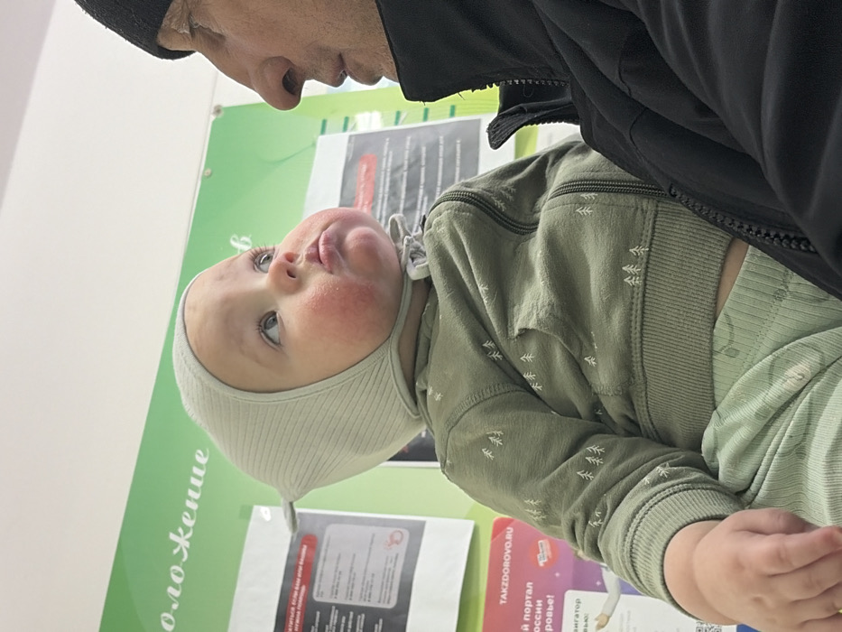 A baby wearing a light green, ribbed bonnet and matching outfit is held by an adult. The baby has rosy cheeks and is looking upwards with a slight pout. The adult, wearing a black jacket and cap, is partially visible at the top of the frame. A light green wall with Russian text is in the background.