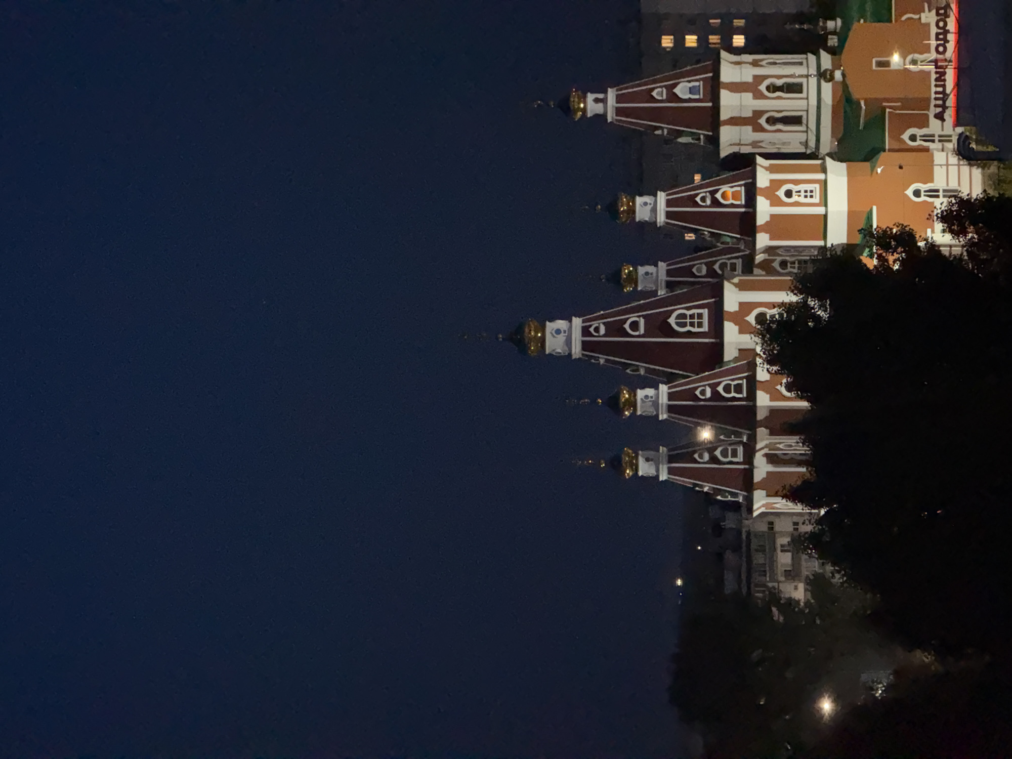 The image depicts the Church of the Nativity of the Blessed Virgin Mary in Otradny, Russia, at night. The church features distinctive red and white onion domes topped with golden crosses. The surrounding area shows dark silhouettes of trees and distant buildings with illuminated windows. A sign with Cyrillic text, partially visible, is present in the lower right corner. The sky is a deep, dark blue, indicating nighttime.