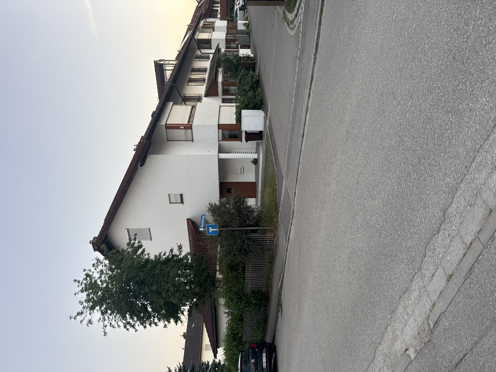 This is a photograph taken in Höhenkirchen-Siegertsbrunn, Germany. The image is oriented vertically, showing a street scene.
The primary subjects are residential buildings and a street. The buildings appear to be townhouses or apartments, with white facades, dark tiled roofs, and multiple windows, many with roller shutters. There are also trees and dense green bushes bordering the properties. A paved street runs alongside the buildings, with a cobblestone curb visible on the right side of the frame. A black car is parked on the side of the road.
A blue and white street sign is mounted on a post, reading "Marcherlweg" with a directional arrow pointing left, and another sign below it indicates a dead end.
The time of day appears to be late afternoon or early evening, given the soft, diffused light and the pale sky with a hint of yellow near the horizon, suggesting sunset or sunrise. The weather seems calm and clear. The scene depicts a quiet residential neighborhood.
The text "Marcherlweg" is visible on the street sign. The license plate of the black car is partially visible, reading "BLBB 3 3907".