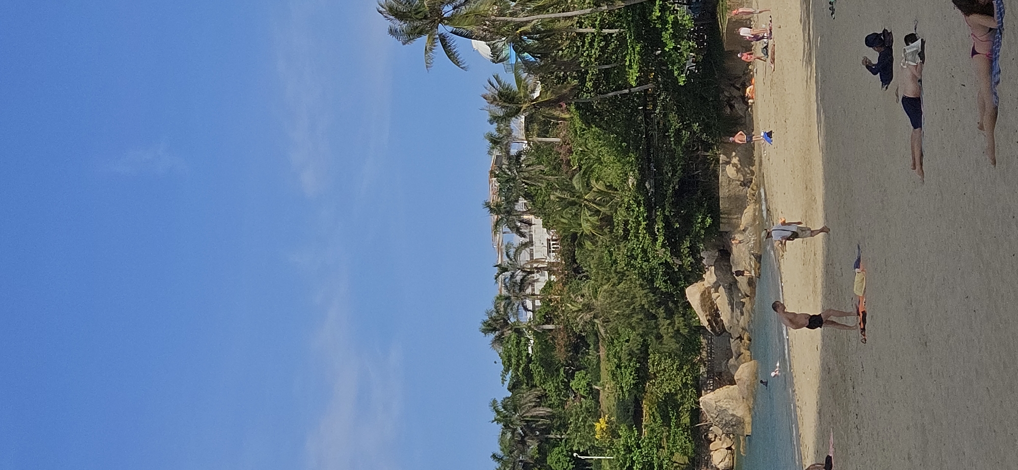 The image depicts a beach scene in Bắc Nha Trang, Vietnam. To the left is a clear, blue sky with few clouds. Lush green vegetation, including palm trees, grows along the slope leading to a building with balconies. On the right, the tan sandy beach is populated with people engaging in various activities such as walking, sunbathing, and standing in the water. Rocks of different sizes are scattered around the edge of the beach. A building can be seen through the trees.