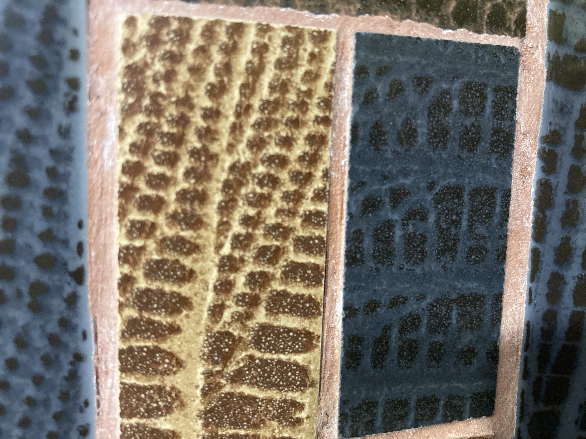 The image displays a close-up of mosaic tiles, likely part of a decorative surface. The tiles are rectangular and framed by thin, reddish-brown grout lines. The upper section features tiles with a textured pattern resembling a honeycomb or overlapping scales in shades of golden brown with darker brown speckles and occasional white dots. The lower section shows tiles with a similar scale-like pattern but in a dark blue or grey hue, also speckled with darker and lighter elements. The overall texture suggests a glossy or semi-gloss finish. There are no people, animals, or specific landmarks visible that would indicate a location in Tyumen, Russia, or any other specific city.