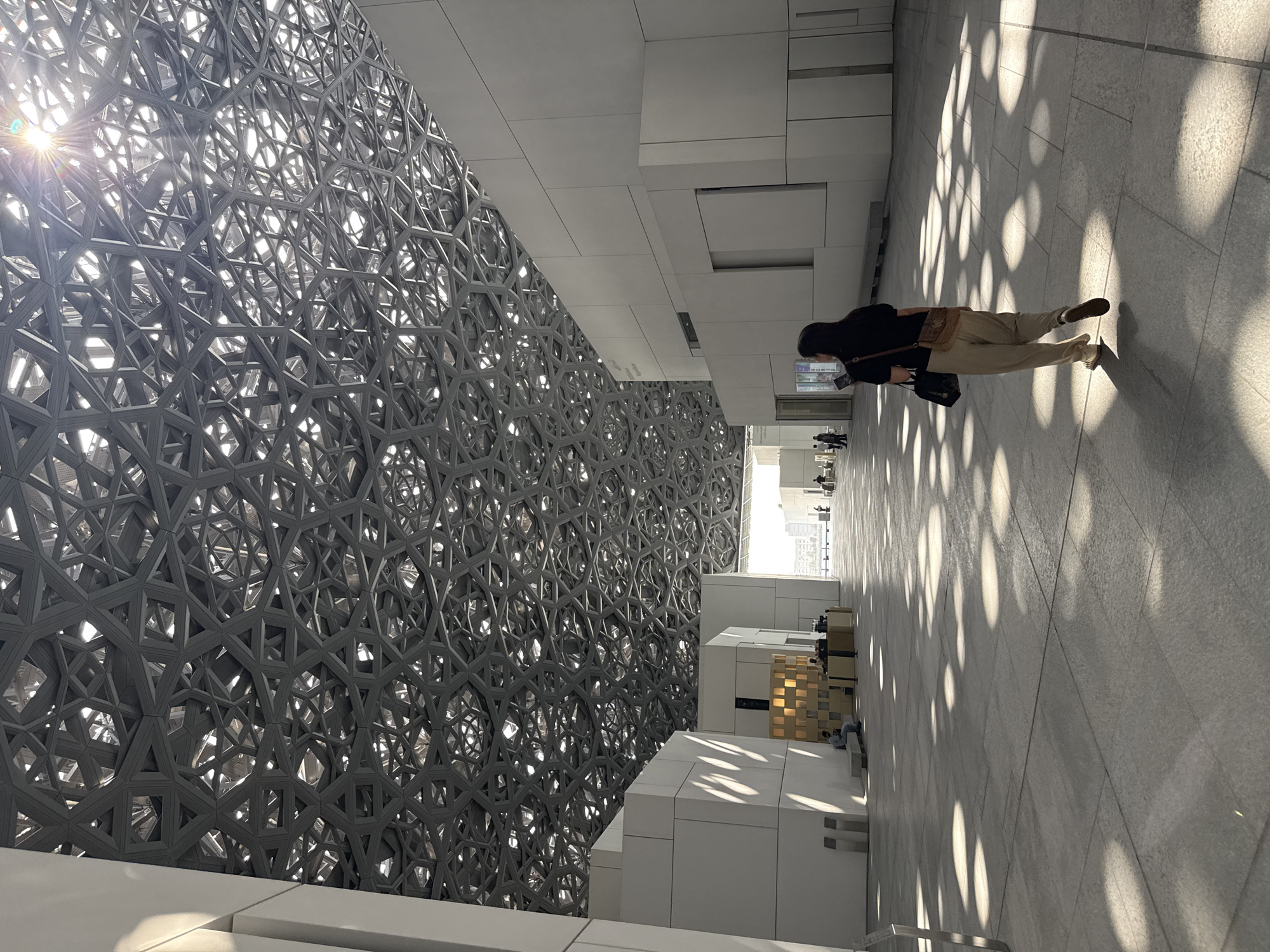 The image captures the interior of the Louvre Abu Dhabi museum in the United Arab Emirates. A woman with dark hair, wearing a black short-sleeved top and light beige pants, walks away from the camera towards a bright, sunlit area. She carries a dark bag over her left shoulder and a light brown crossbody bag. Her left foot is mid-stride, suggesting she is walking at a moderate pace. The floor is paved with large, light-colored tiles, and the sunlight filters through an intricate, geometric lattice-like roof structure, casting circular patterns of light and shadow across the pathway. White, angular architectural elements form the walls of the museum, with some areas featuring darker paneling. In the background, other visitors can be seen in the distance, and a glimpse of buildings and possibly water is visible beyond the museum's interior. The overall impression is one of modern architecture and diffused natural light.