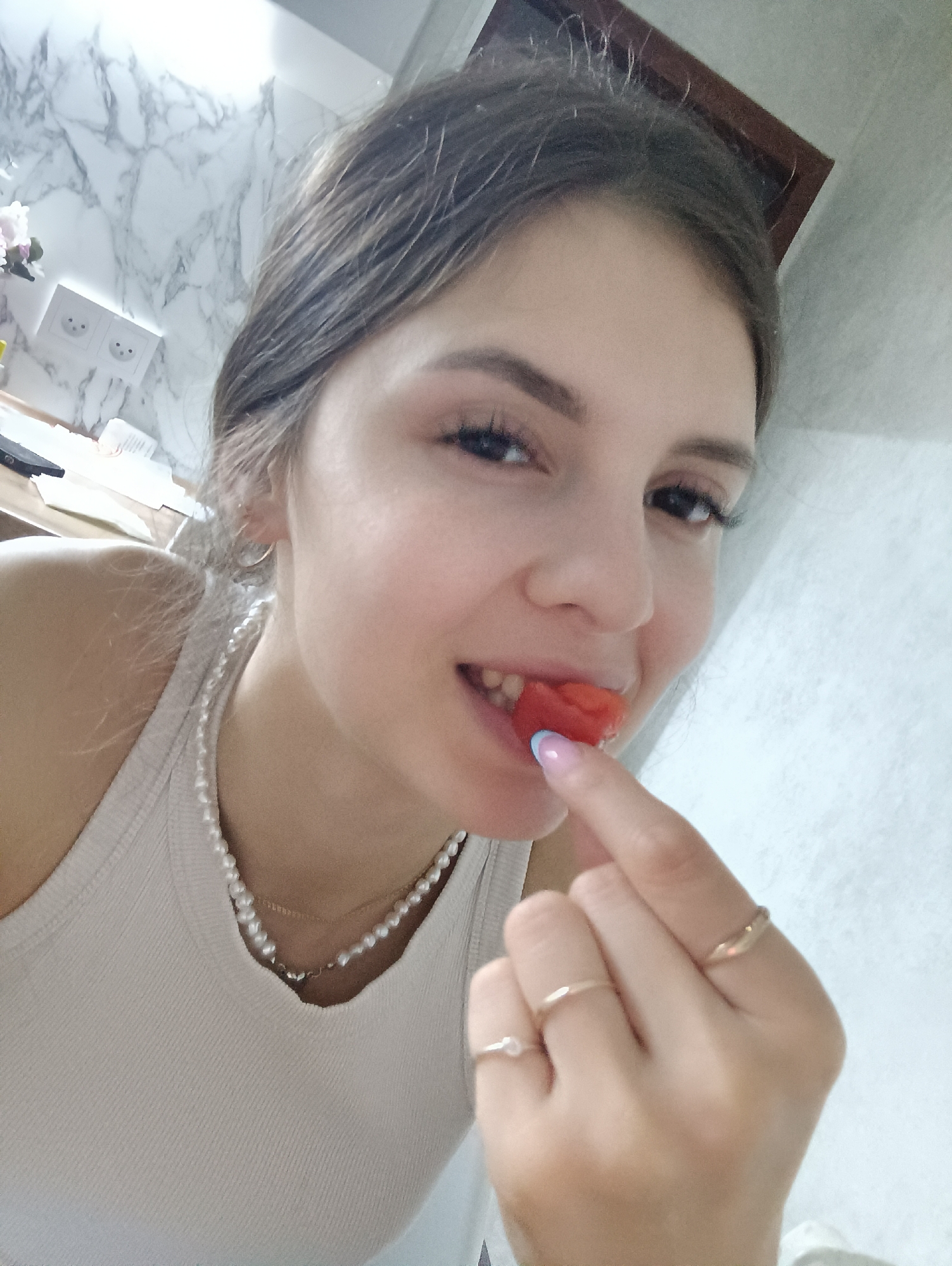 A young woman with dark hair is holding a piece of red food, possibly watermelon, to her mouth with her left hand. She is wearing a white ribbed tank top and a double-stranded pearl necklace, with a delicate gold necklace layered underneath. She has three gold rings on her fingers, and her fingernail is painted with a pale blue tip. She is inside with a marble-patterned wall behind her to the left, and a white wall to the right. There is an electrical outlet on the wall behind her and a picture frame towards the top right. The location is not specified, but the question states it is Chaikovsky, Russia.