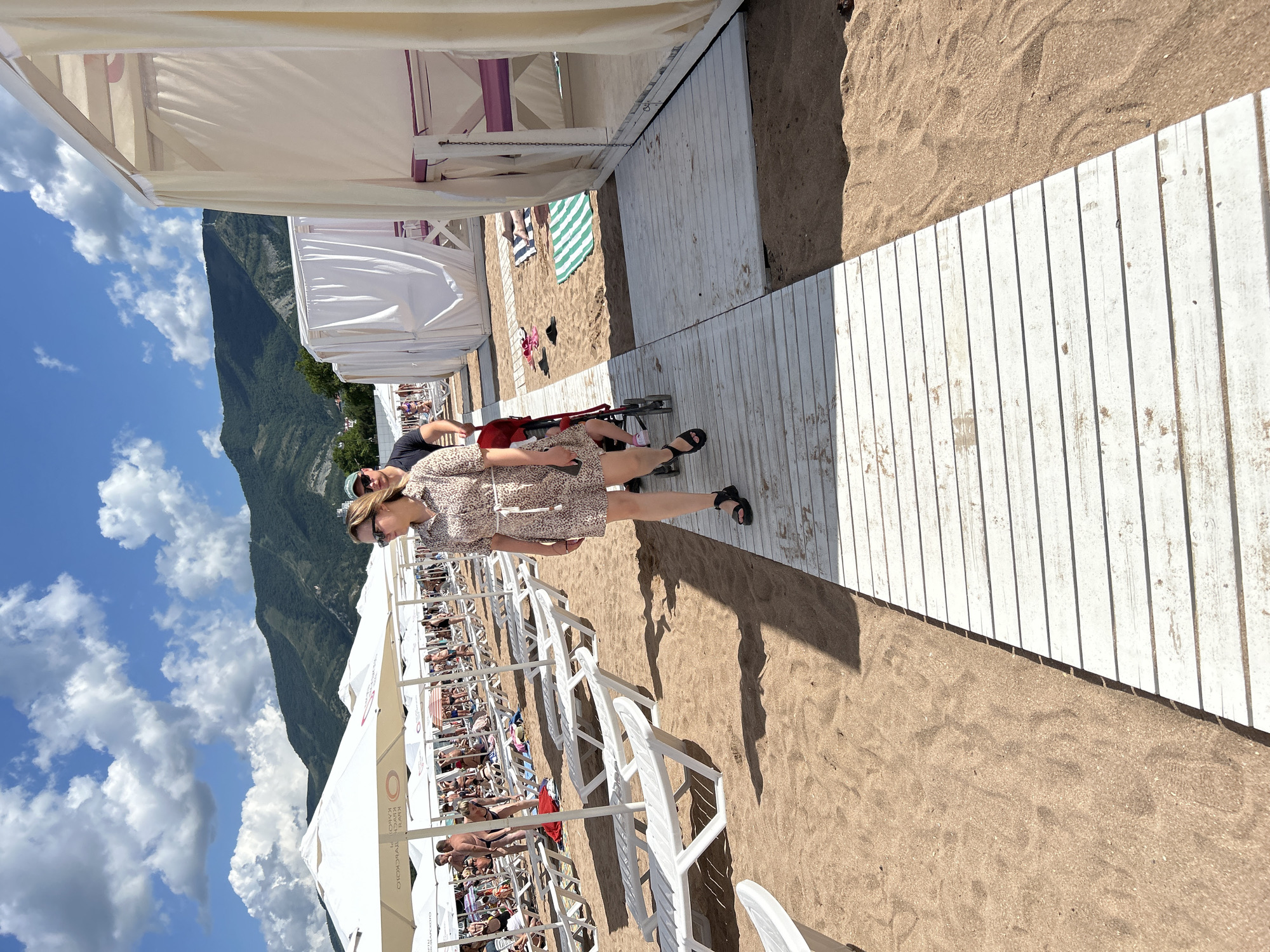 On a sunny day at the beach in Gelendzhik, Russia, a woman in a light-colored, patterned dress walks along a white wooden boardwalk. She is wearing sunglasses and black sandals, and her hair is blonde. Behind her, a man in a black t-shirt and a green baseball cap pushes a stroller with a child inside. The beach is lined with white lounge chairs and umbrellas, and in the background, lush green mountains rise against a bright blue sky with scattered clouds. White cabanas with draped curtains are also visible along the side of the boardwalk. The sand is a warm, light brown, and footprints are visible on the boardwalk.