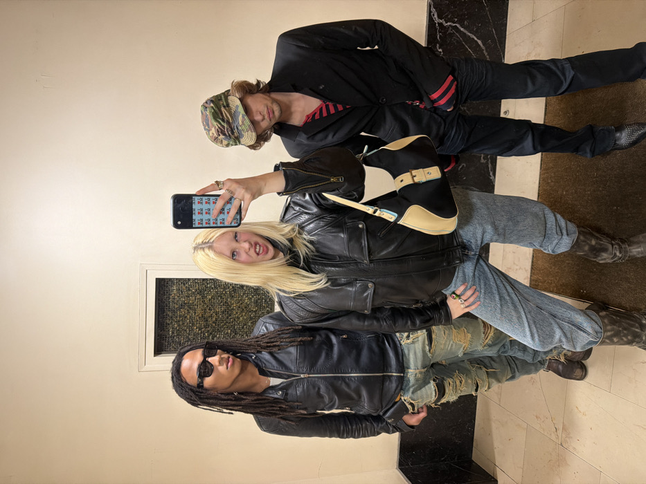 Three individuals are depicted in an indoor setting, identified as Paris, France. A light-skinned female in the center, with long blonde hair and multiple rings on her fingers, holds a black smartphone with a patterned light blue case in her raised right hand, appearing to capture a mirror selfie. She wears a black leather jacket, light blue jeans, dark boots, and carries a black and beige shoulder bag. Her facial expression is characterized by a scrunched nose and open mouth, revealing teeth. To her left stands a dark-skinned male with dreadlocks, wearing a black leather jacket, rectangular sunglasses, heavily distressed light blue jeans, and dark boots. He looks towards the right. To the female's right stands a light-skinned male wearing a camouflage bucket hat, a black blazer over a red and black striped shirt, dark trousers, and dark pointed shoes. He looks slightly downward. The background consists of plain, light-colored walls featuring a rectangular recessed panel with a dark, textured infill. The floor is composed of a light-colored tiled section with a dark horizontal border, transitioning to a darker, textured mat in the immediate foreground.