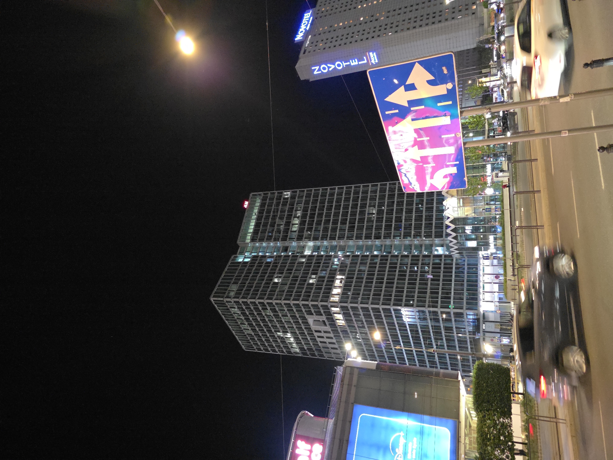 The image shows a night scene in Warsaw, Poland, with several illuminated buildings visible against a dark sky. The Novotel Warsaw Centrum hotel is prominent, with its sign illuminated in blue. A large, modern office building with a grid-like façade is also visible. A street sign with directional arrows stands near the sidewalk. Cars are visible on the roadway, with motion blur indicating movement. Streetlights are illuminating the scene.