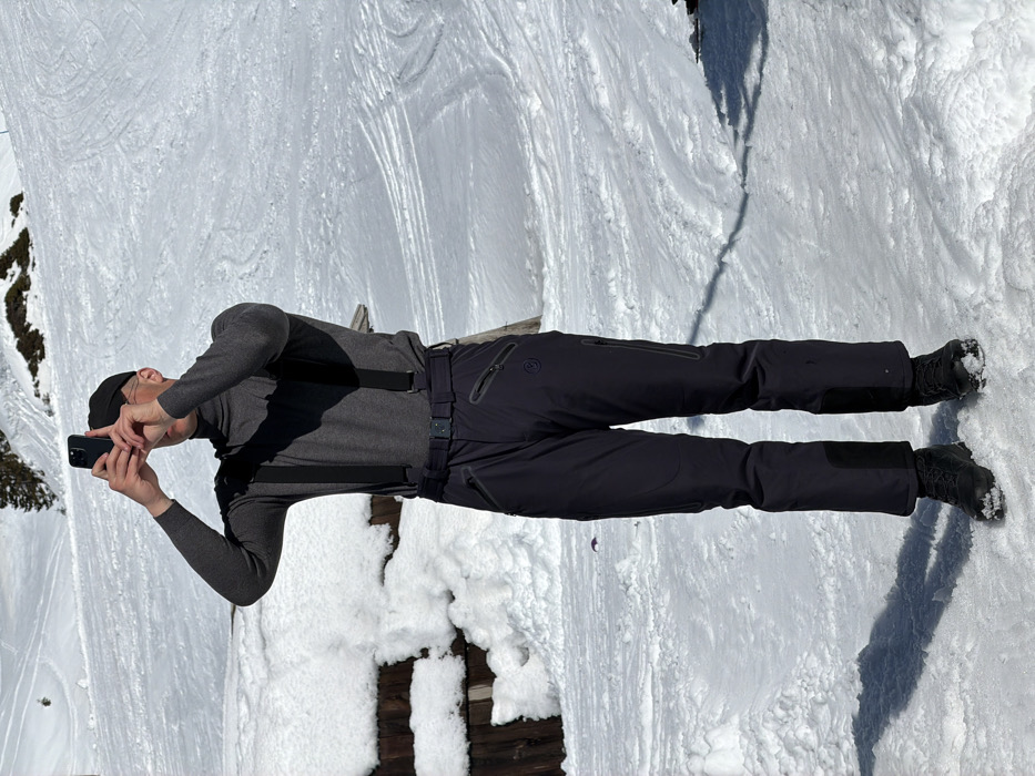 An adult male stands in a snow-covered outdoor environment in Vranjak, Montenegro. He is positioned centrally, facing slightly right of the camera. He wears a dark grey long-sleeved turtleneck shirt with black suspenders, dark grey or black snow pants featuring zippered pockets and a logo on the upper right thigh, a black belt with a metallic buckle, and black boots dusted with snow. A dark baseball cap covers his head. His arms are raised, holding a dark-colored smartphone horizontally with both hands, obscuring most of his face as he appears to be taking a photograph or video. The ground is entirely covered in white snow, displaying various tracks. To the left, a wooden structure is partially covered by a thick layer of snow. In the background, a snow-covered hillside with distinct ski tracks slopes upwards, featuring scattered trees or brush. The scene is illuminated by direct sunlight, casting shadows on the snow.