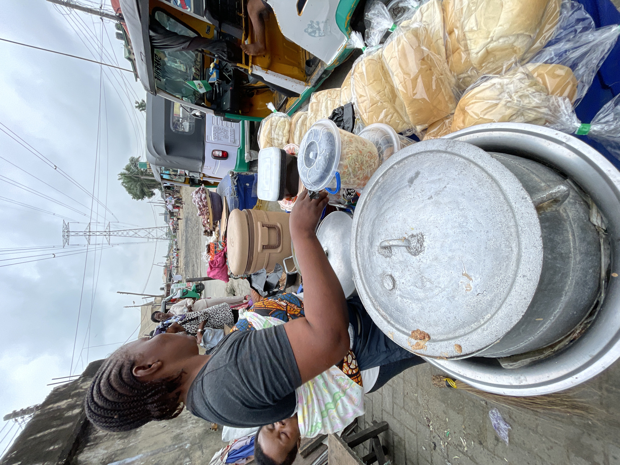 A Black woman with braided hair is standing in what appears to be a busy street market in Ado, Nigeria, with a baby wrapped in a colorful cloth carrier on her chest. She is wearing a dark t-shirt. She is attending to large metal cooking pots on a table in front of her. Behind her, several other people are visible, including a woman in a patterned dress and a man in a white shirt and dark pants near a tuk-tuk. The environment suggests an outdoor market with various goods for sale, including bread wrapped in plastic and containers of food. Power lines are visible in the sky, indicating an urban setting.