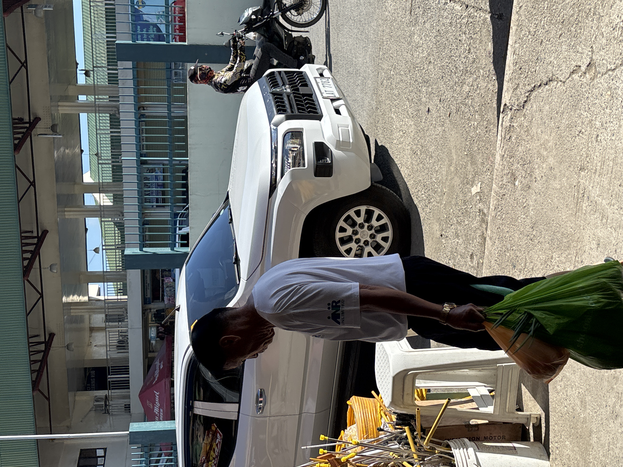 A man in a white t-shirt and dark pants walks in front of a white SUV and a motorcycle with a rider. The man is carrying a green plastic bag in his right hand. The motorcycle rider is wearing a helmet and a patterned shirt. In the background, there is a building with a green roof and metal fencing. The scene appears to be outdoors, possibly on a street or sidewalk. The license plate of the SUV is visible and reads "LAT 7782". The image was likely taken in Tagum City, Philippines, given the context provided.