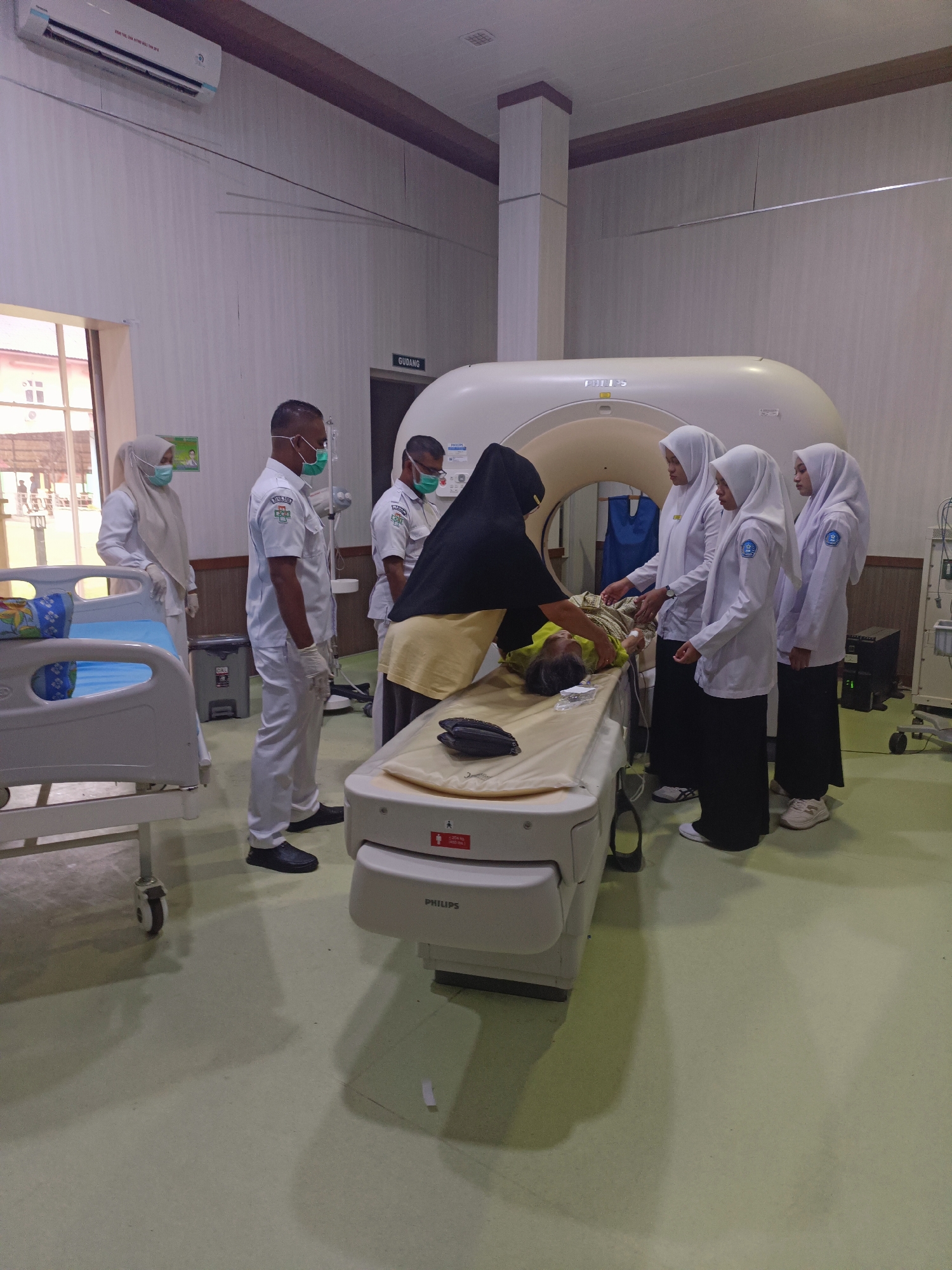 In a medical imaging room, likely a CT scan suite, at the Mesjid Reuntoh health clinic in Indonesia, a young patient lies supine on a movable examination table, positioned in front of a large, white CT scanner. The patient, appearing to be a child or adolescent, is wearing a light green gown. A woman in a black headscarf and yellow tunic, presumably a medical professional, is attending to the patient, adjusting a blanket or sheet.
Standing to the right of the scanner are three young women in white lab coats and hijabs, likely medical students or junior staff, observing the procedure. To the left of the CT scanner, two men in white medical uniforms and masks are also present, one with a name tag reading "FARIADI", and both appear to be supervising or assisting. Another individual in a white hijab and uniform stands further to the left, near a medical trolley with a blue mattress.
The room is brightly lit with fluorescent lighting from an air conditioning unit mounted high on the wall. The walls are a neutral color, and the floor is a pale green linoleum. A doorway labeled "GUDANG" (Storage) is visible in the background. Medical equipment, including a cart with a screen and various wires, is positioned to the right of the scanner. The overall atmosphere suggests a clinical examination or training scenario.
