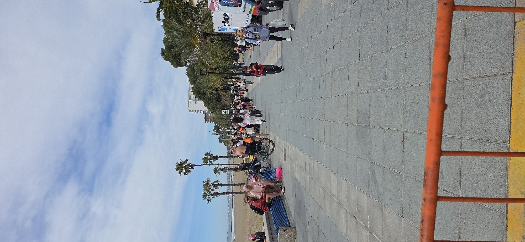 The image depicts a street scene in Benicàssim, Spain, with a view of the coast on a day with a partially cloudy sky. Palm trees line the street alongside buildings in the background. A group of people appears to be walking or gathering on the sidewalk. A few people are seated on benches near the beach. A bicycle is parked near the sidewalk. An orange barrier is visible in the foreground. An ambulance is parked on the side of the road further ahead. The sidewalk is paved with gray tiles.