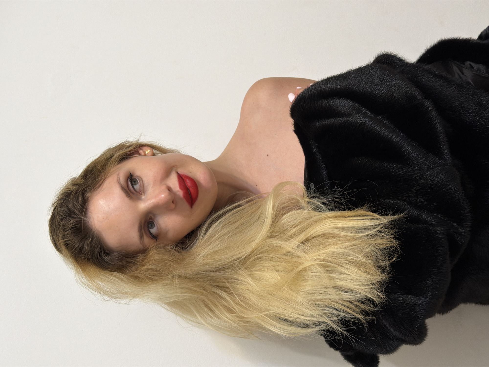 A woman with blonde hair is lying down against a white background, wearing a black fur coat. Her head is tilted towards the camera, and she is wearing red lipstick. There is no indication of the location being in Budva, Montenegro.