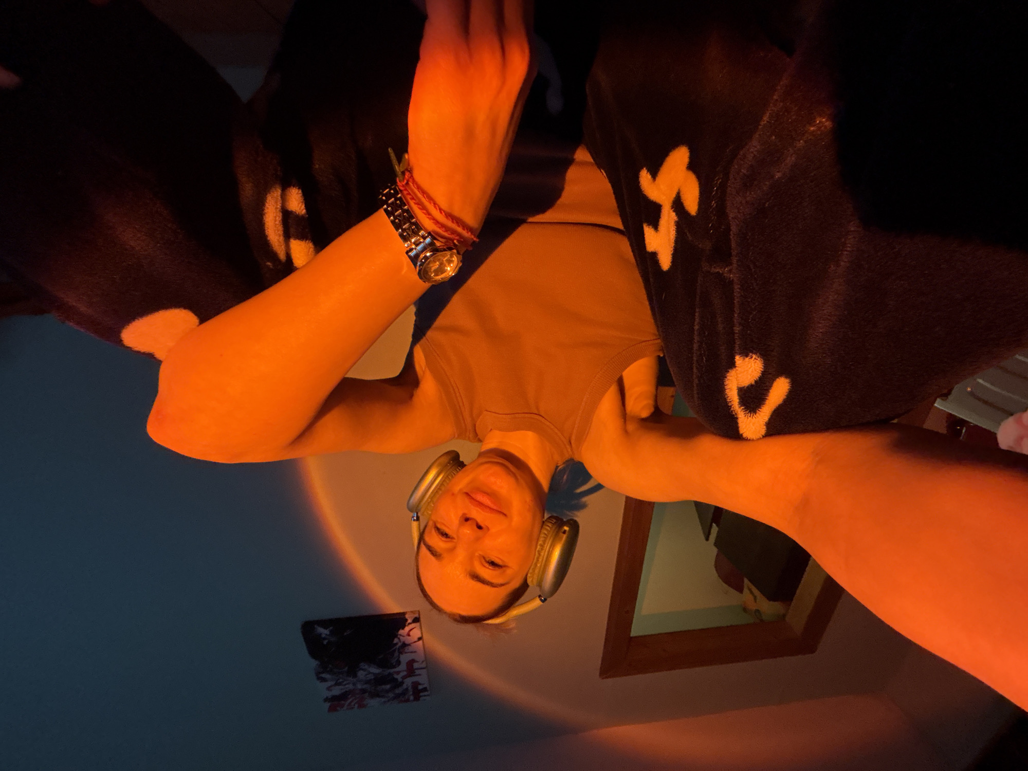 A person is upside down in an interior space. They are wearing headphones and a tank top, and a watch and bracelets on their wrist. The person is positioned in front of a wall decorated with an artwork and a wooden-framed picture. The person is surrounded by a dark fabric with light-colored symbols. A warm orange light is projected onto the wall and the person. The location is Unknown, Russia.