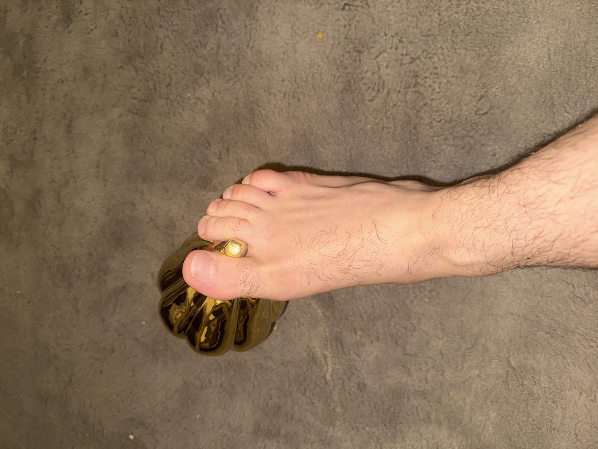 A bare foot rests atop a golden, ornamental pumpkin on a gray carpeted surface. The foot is positioned with the toes pointing towards the pumpkin's top, where the great toe presses down. A ring is visible on the great toe. The location is unknown, but the request specified Blagoevgrad, Bulgaria.