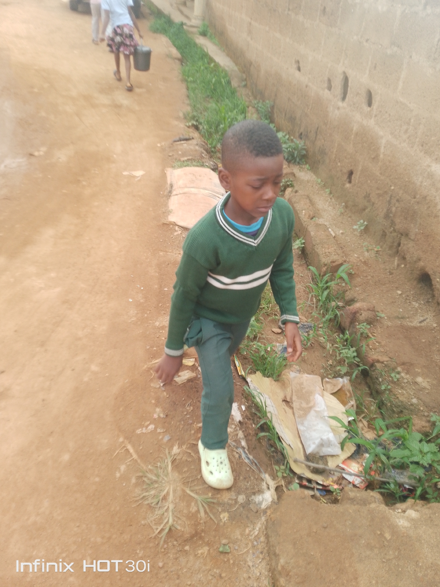 A young boy, appearing to be of school age, walks on a dirt path next to a shallow, overgrown drainage ditch in Jos, Nigeria. He is wearing a green V-neck sweater with white stripes on the chest and sleeves, green pants, and white perforated clogs. His left hand is extended as if reaching for something or steadying himself. In the background, another person, possibly a girl in a skirt, is walking away from the camera, carrying a black bucket in their right hand. The environment consists of a dirt road, the drainage ditch with visible vegetation and debris, and a rough brick wall on the right side of the frame. The overall impression is of a scene depicting everyday life in a less developed urban area.