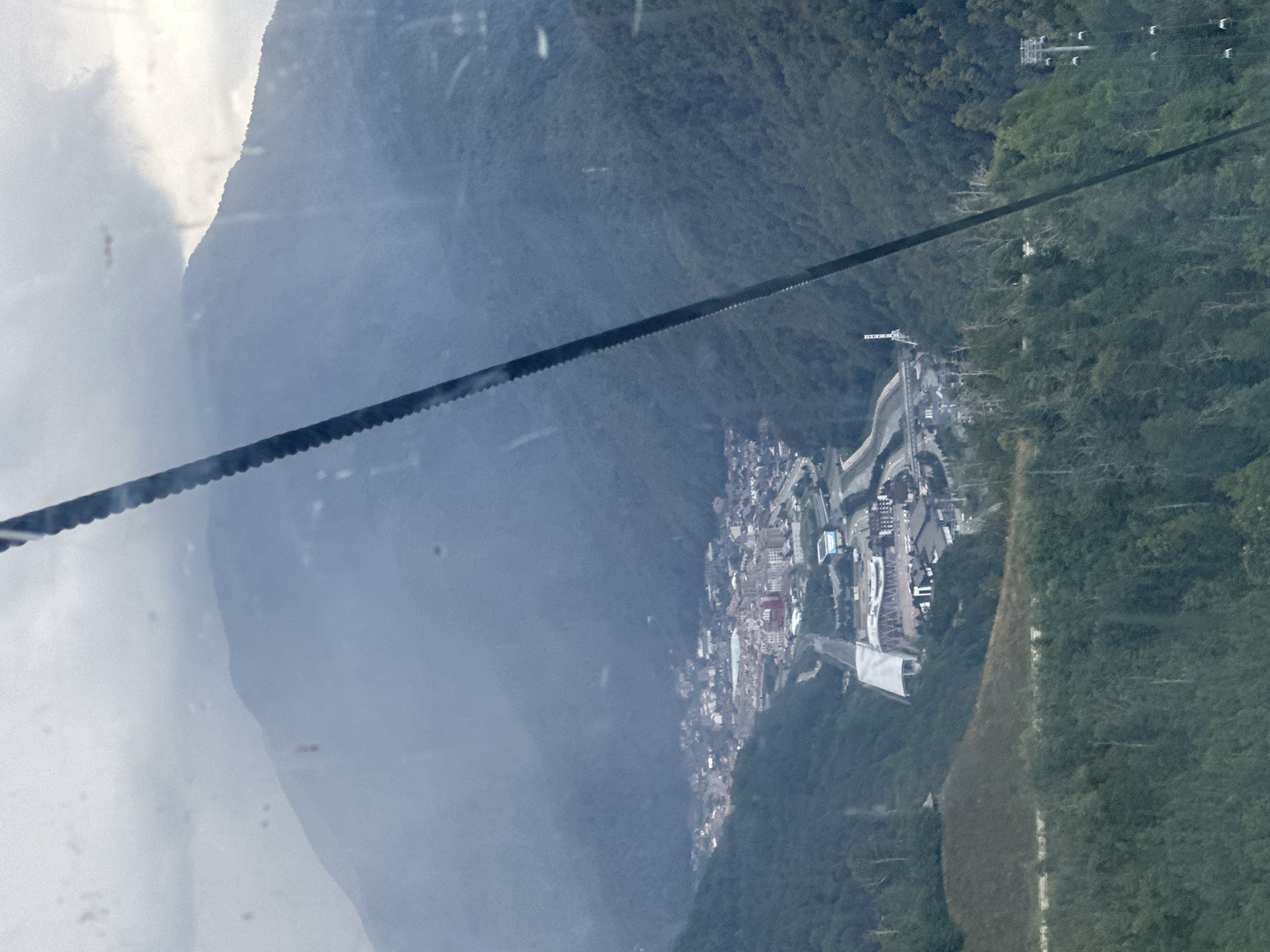 The image presents a panoramic view of Estosadok, Russia, a mountain resort town nestled within a valley. The scene is captured from an elevated vantage point, likely from a cable car, as indicated by a thick cable bisecting the frame diagonally. Lush green mountains, densely covered with trees, form the backdrop and flank the valley. A prominent cable car line with multiple cabins descends along the right side of the frame, disappearing into the dense foliage.
In the valley, a cluster of buildings forms the town of Estosadok. The architecture suggests a modern development, with various structures of different sizes and colors. A notable building with a large, arched roof structure, possibly a sports arena or stadium, is visible near the center of the town. Roads and pathways are interspersed throughout the urban area, connecting the various buildings. The overall atmosphere is that of a serene, mountainous landscape dotted with human settlements and infrastructure. The sky is overcast with clouds, casting a soft, diffused light over the scene.