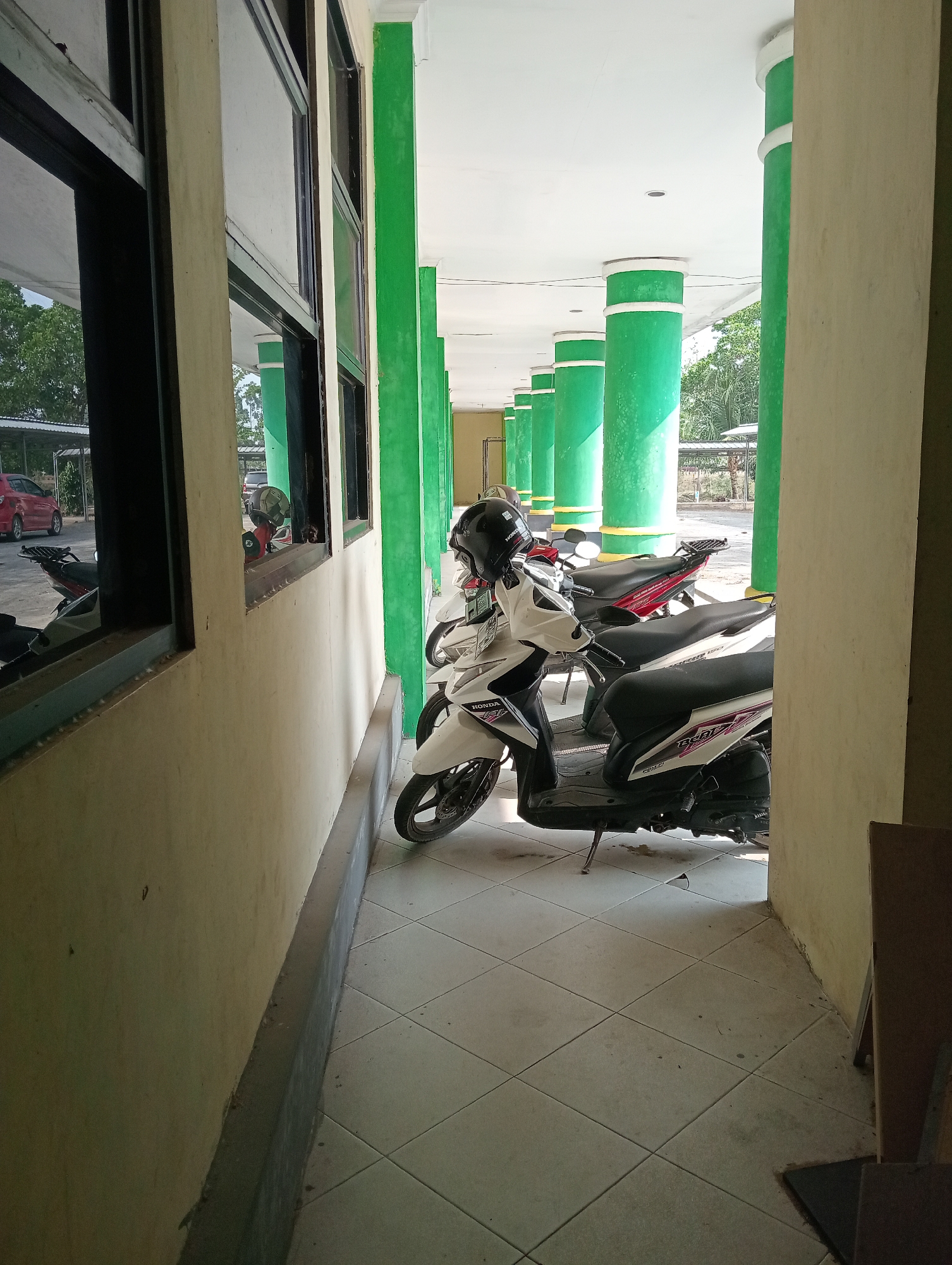 The image depicts an outdoor hallway with a tiled floor and green pillars supporting a white roof. Several motorbikes are parked in the hallway. One white motorbike with a black helmet on its seat is prominently featured in the foreground. In the background, a red car is visible under a canopy. The scene appears to be a public space, possibly a school or office building, given the architecture and the presence of parked vehicles. The lighting suggests it is daytime. The overall impression is one of a typical urban or suburban scene in Indonesia.