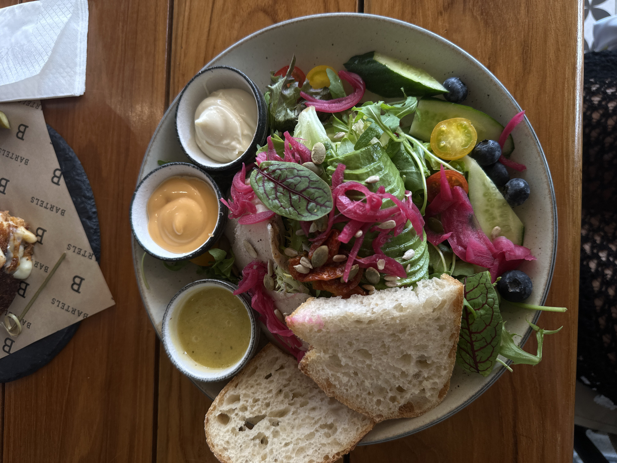 A top-down shot shows a plate with a salad and condiments on a wooden table. The salad contains greens, avocado, tomatoes, cucumbers, blueberries, pickled onions, and seeds, with bread on the side. There are three small bowls with white, orange, and yellow sauces. A coaster with the name "Bartels" is visible, along with a napkin and a dessert item. The setting appears to be a cafe or restaurant. The location is Choeng Thale, Thailand, based on the brand of the coaster. A person is visible at the right edge of the frame, wearing a lacy or patterned fabric.