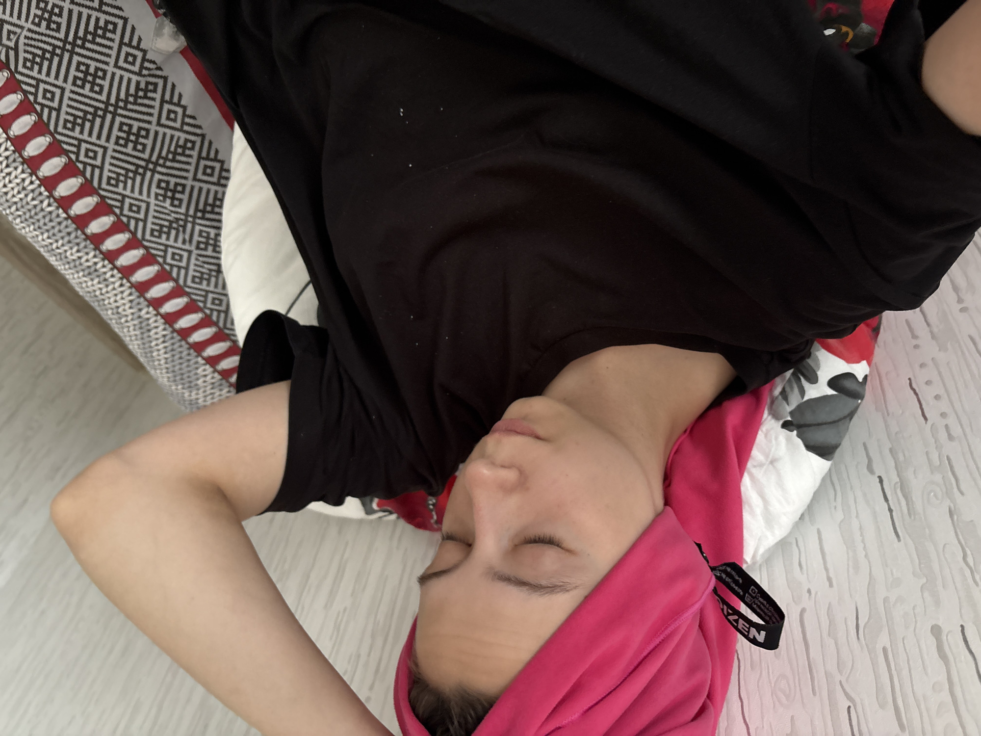 A woman lies in an indoor setting with a pink towel wrapped around her head. She is wearing a black t-shirt and rests on a patterned pillow. The floor is a light, possibly faux wood material. The exact location within Perm, Russia is not discernible from the image.
