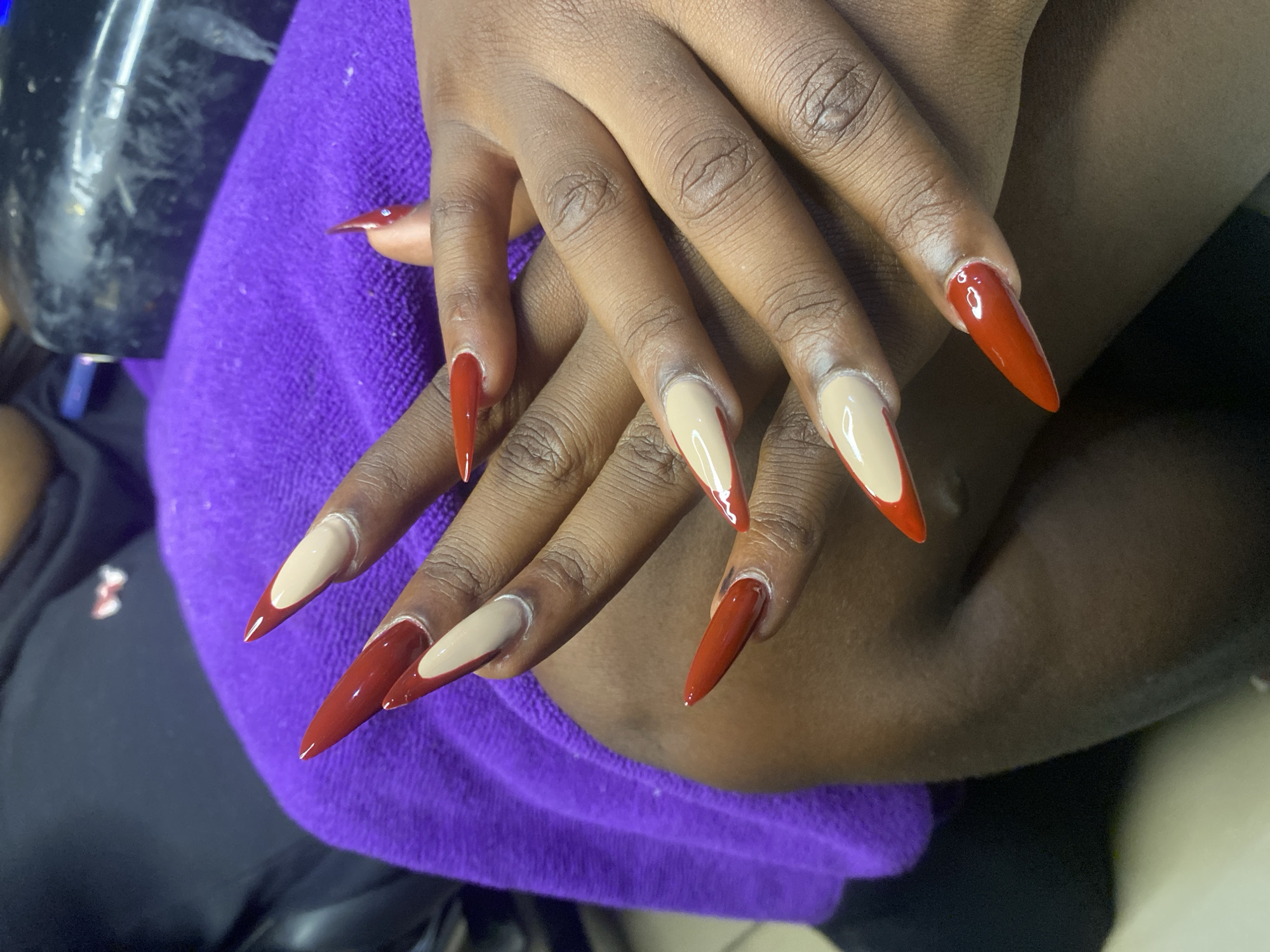 A pair of hands with long, stiletto-shaped acrylic nails, painted in a dual-tone design of deep red and nude, rests on a purple towel. The nails are neatly shaped and glossy. The hands appear to belong to a person of African descent. The background is slightly blurred but suggests an indoor setting, possibly a salon, with a black piece of equipment and a hint of a dark garment. The location is identified as Kubwa, Nigeria.