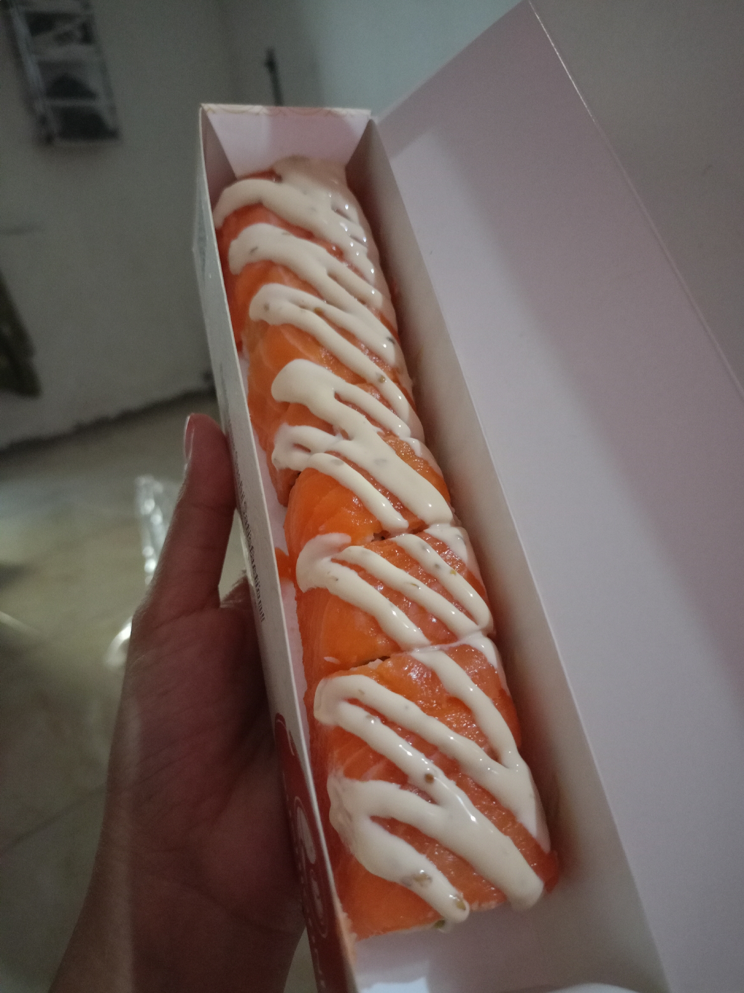 A hand is holding a white cardboard box containing several pieces of salmon sushi rolls. The sushi rolls are topped with a creamy white sauce drizzled in a zig-zag pattern. The background appears to be an indoor environment with a blurry grey floor and wall. The scene does not provide any contextual information related to the city of Pasir Gadung, Indonesia.