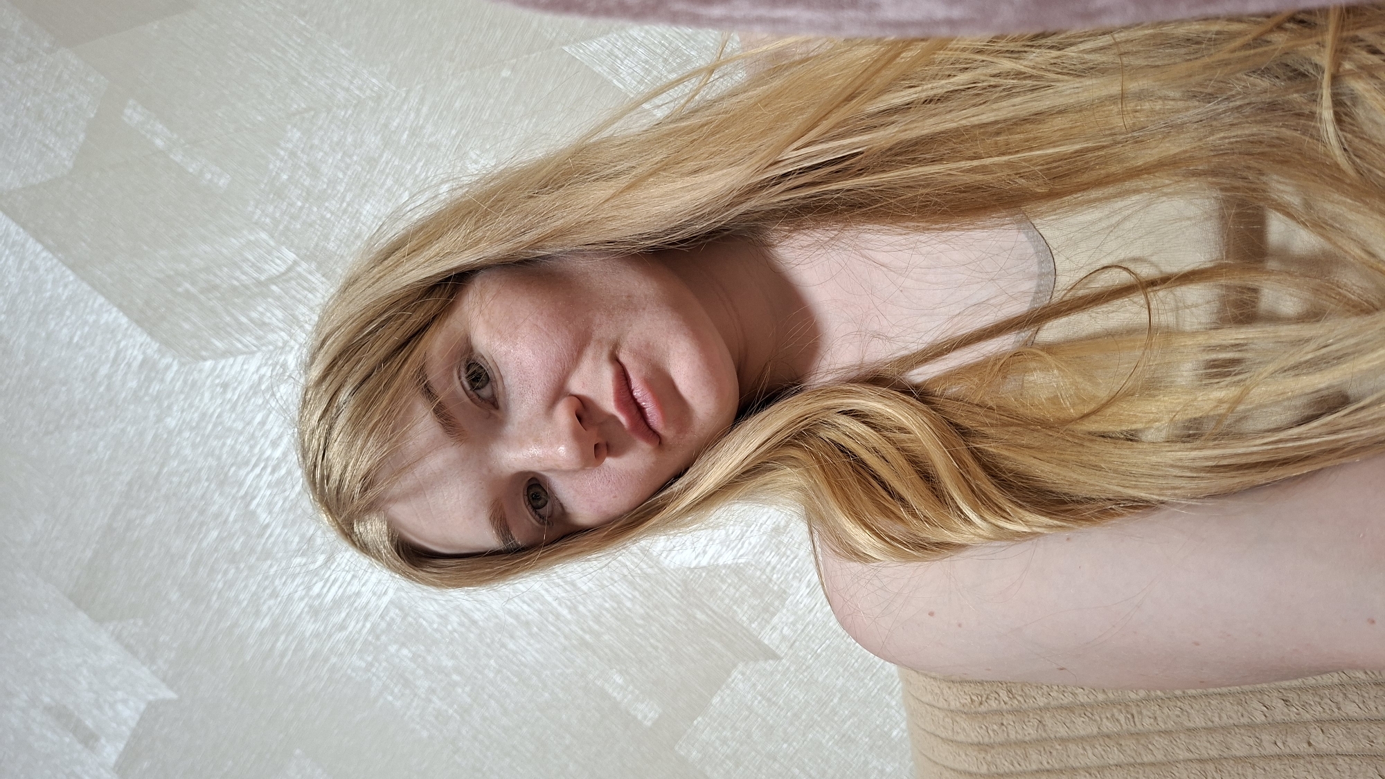 A young woman with fair skin and long, blonde hair is the primary subject of this close-up, slightly tilted shot. Her hair is styled with loose waves, cascading around her face and shoulders. She has light-colored eyes and a neutral expression.
The background is a textured wall with a subtle, patterned design. In the top part of the image, a section of a pinkish blanket is also visible. The presence of an arm suggests she may be lying down on a beige/light brown fabric surface.
The location of the image cannot be identified from the given information. It may or may not be in the city of Большой Букор, Russia.