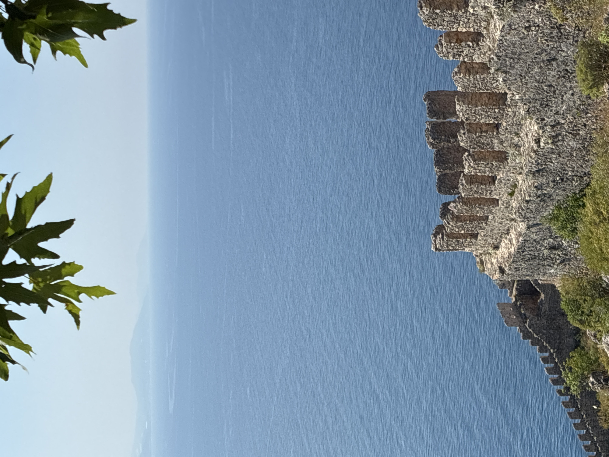 The image depicts a scenic view of the Alanya Castle, a historical landmark located in Alanya, Turkey. The castle walls, constructed from rough-hewn stones, are visible in the foreground, with battlements clearly defined along the ramparts. The ancient fortress overlooks the vast expanse of the Mediterranean Sea, which dominates the middle ground of the image with its deep blue, rippling surface. In the distance, hazy landmasses or islands are faintly visible on the horizon, adding depth to the landscape. The upper portion of the image features lush green leaves from a tree, framing the scene and creating a natural border. The sky is a clear, pale blue, suggesting a bright, sunny day. The overall impression is one of historical grandeur meeting natural beauty.