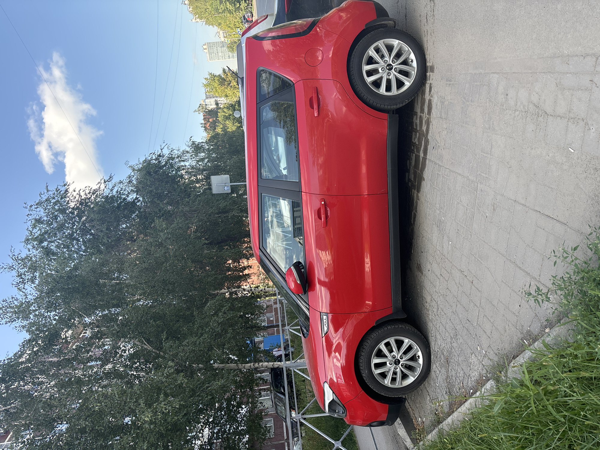 A bright red Kia Soul is parked on a cobblestone sidewalk next to a street in Novosibirsk, Russia. The car is positioned parallel to the street, with its passenger side facing the camera. The sidewalk is bordered by a metal fence and green grass. Behind the fence, a multi-story residential building with light-colored brick and windows is visible, partially obscured by the dense foliage of tall, leafy trees that line the street. The sky is blue with scattered white clouds. In the background, other buildings and trees can be seen. The scene appears to be a typical urban environment on a clear day.