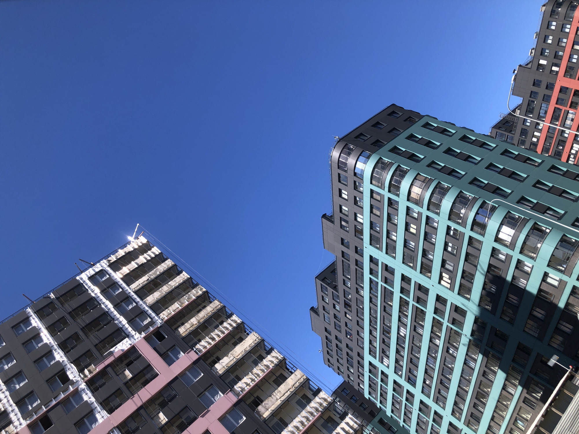 This image presents a low-angle, upward-looking view of several modern residential buildings against a clear, vibrant blue sky in Saint Petersburg, Russia.
In the upper left quadrant, a tall multi-story building is visible, likely under construction or renovation. Its facade predominantly features dark grey cladding, with vertical sections accented in a light pinkish-mauve hue. Multiple balconies are present, many appearing unfinished with exposed concrete or white insulating material. A prominent sun glare or reflection is visible near the top right corner of this structure.
Dominating the lower-center and right portions of the frame are two other multi-story residential buildings, characterized by their contemporary design and distinct color blocking. The primary building in the foreground features a dark grey upper section that transitions to a prominent teal or turquoise color across the majority of its facade. This building incorporates numerous windows and features smooth, curved architectural elements, particularly at its corners.
Partially visible to the right and slightly behind the teal-colored building is another similar structure, which also employs dark grey and a distinct red-orange color block on its facade. The bright daylight creates strong contrasts and reflections on the windows.
No individuals, vehicles, or specific street-level identifiers are present within the frame to pinpoint an exact address or specific activity. The scene solely focuses on the architectural forms and their interaction with the bright, cloudless sky.