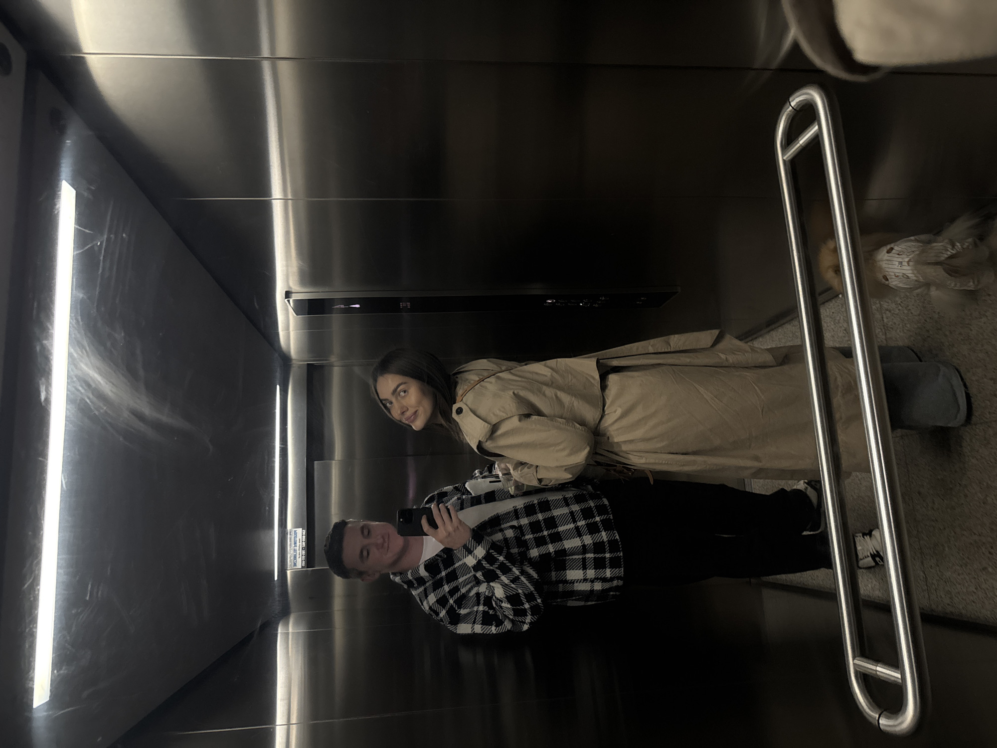 A couple takes a selfie in an elevator with stainless steel walls. The man, wearing a black and white plaid shirt, holds a black smartphone. The woman, standing behind him, wears a beige trench coat. A silver grab bar is visible in the corner. A dog is present in the elevator. The location appears to be in Warsaw, Poland based on elevator signage.