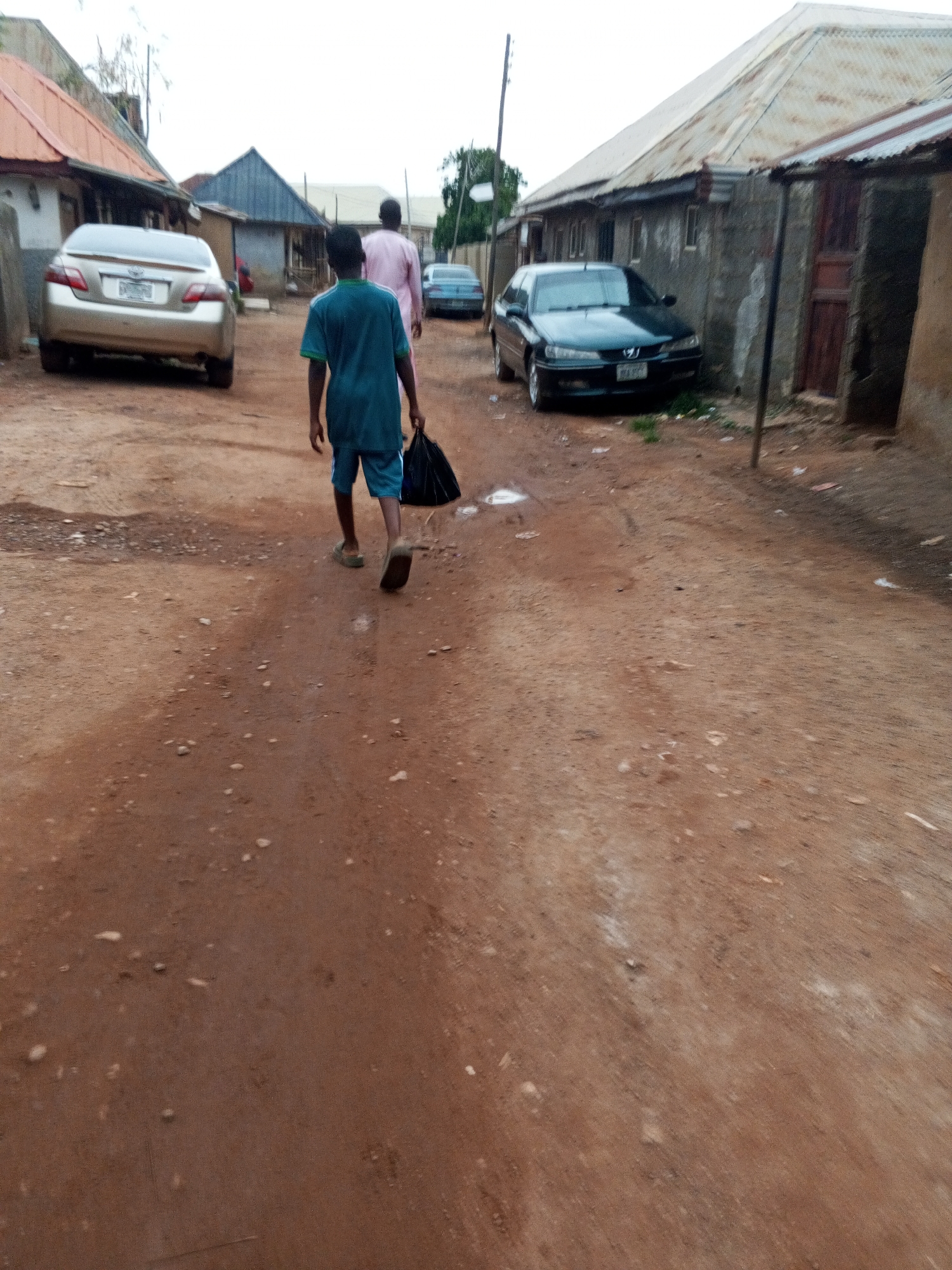 The scene is a dusty, unpaved street in what appears to be a residential area. The location is likely in Abuja, Nigeria.
A child wearing a green shirt and shorts is walking away from the viewer, carrying a black plastic bag. An adult wearing a pink robe-like garment is walking a short distance ahead of the child.
Two cars are visible: a gold Toyota Camry parked on the left side of the street, and a dark green Peugeot sedan parked on the right.
Buildings with corrugated metal roofs line the street. The buildings appear to be made of concrete blocks. One building has a wooden door.