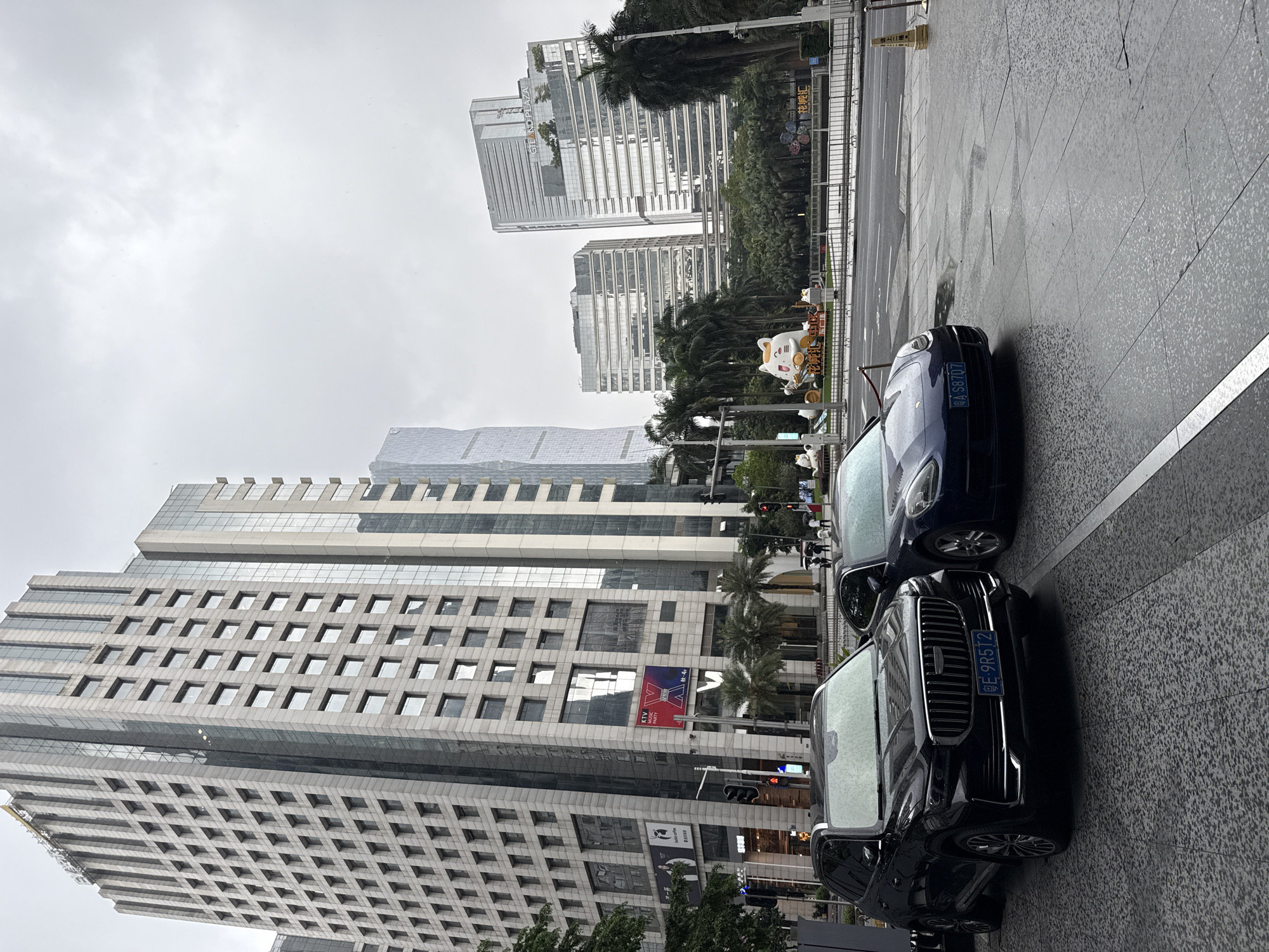 An overcast day in Tianhe District, China, features a street scene with modern architecture and vehicles. In the foreground, a dark-colored SUV with license plate "GE-9R5T2" is parked on a wet, tiled sidewalk. To its right, a dark blue Porsche Panamera with license plate "A-S8707" is also parked. Behind the vehicles, a street with traffic lights is visible. Further back, a line of palm trees and lush greenery separates the street from a plaza area. In the plaza, a large, cartoonish white cat sculpture with a gold halo is prominently displayed, surrounded by other smaller decorations and what appears to be a flower arrangement. Beyond the plaza, several multi-story glass-fronted buildings dominate the skyline, with one in the mid-ground bearing the sign "GRAND PLAZA." The sky is a uniform gray, suggesting cloudy or rainy weather. Several individuals with umbrellas are visible walking in the background, further indicating inclement weather. The overall impression is an urban environment, likely a commercial or business district, during a period of precipitation.