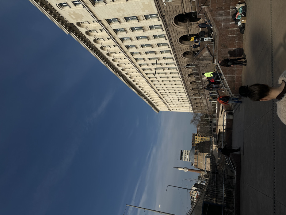 The image depicts a daytime scene in Sofia, Bulgaria, specifically the Largo architectural complex. The view is dominated by the Council of Ministers building on the right, a large, multi-story structure with a light-colored stone facade, numerous rectangular windows, and a lower level featuring a dark stone base with a series of prominent arched passageways. Multiple sets of wide, red-brown granite staircases lead up to the building's entrance.
In the middle ground, towards the left, the Banya Bashi Mosque is visible, characterized by its brick base, large dome, and a slender, light-colored minaret with a pointed cap. Adjacent to the mosque, a sunken archaeological site, identifiable as part of the Serdica complex, features visible ancient stone foundations and structures, with dark metal railings surrounding it. Various other urban buildings are visible in the background on the left.
The sky is a clear, deep blue with minimal, wispy white cloud formations. Bright sunlight illuminates the upper portion of the main building and casts long shadows across the foreground plaza and the left side of the scene, indicating a low sun angle. Leafless trees are scattered throughout the background, suggesting a cooler season.
Multiple individuals are present. In the immediate foreground, facing away from the viewer, is a person with dark hair tied in a bun, wearing a light-colored, textured outer garment over a dark top. Approximately 10-15 people are observed on and around the granite staircases leading to the Council of Ministers building; one individual wears a bright neon yellow/green jacket, and another wears a red/orange jacket, both ascending stairs. Other individuals are dressed in dark or muted clothing, walking or standing. In the sunken archaeological area on the left, a solitary dark-clad figure is visible. Near the base of the right staircase, a small collection of black plastic bags and a cardboard box are present on the ground. Modern streetlights are visible on the left side of the image.