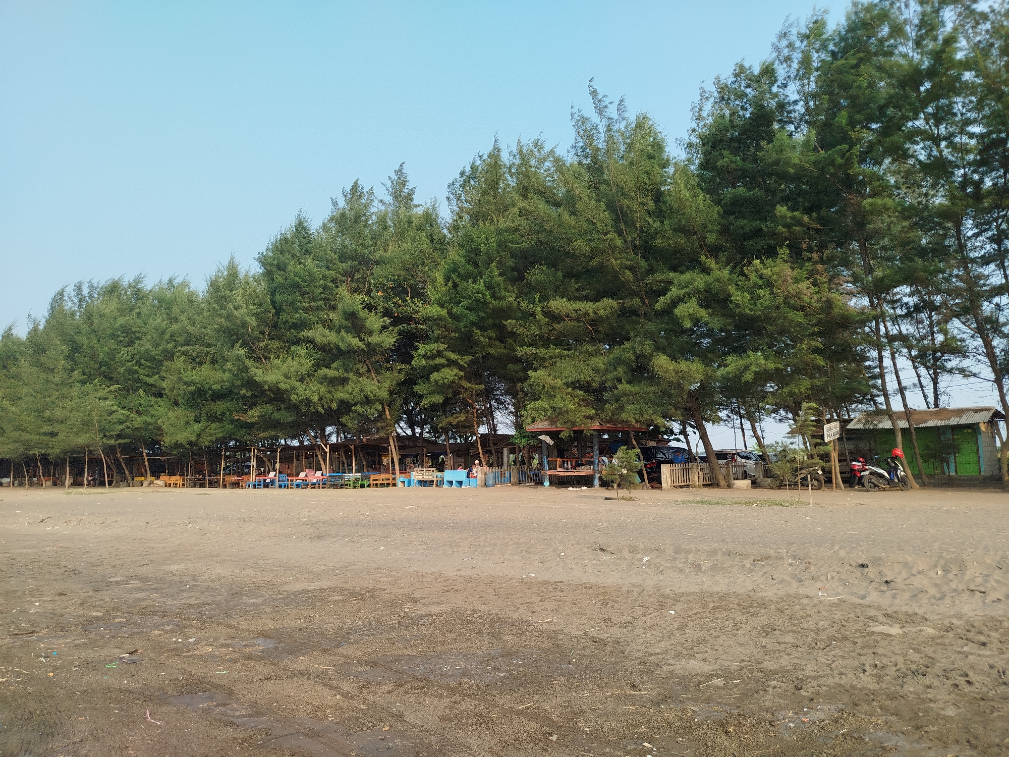 The image shows a sandy beach with a line of trees in the background. Several small structures, likely food stalls or shelters, are visible behind the trees, with tables and chairs set up outside some of them. A few people are seated at the tables. A motorcycle with two helmets is parked on the right side of the image, next to a small green building. The sky is clear and blue. The location is identified as Jungsemi, Indonesia.