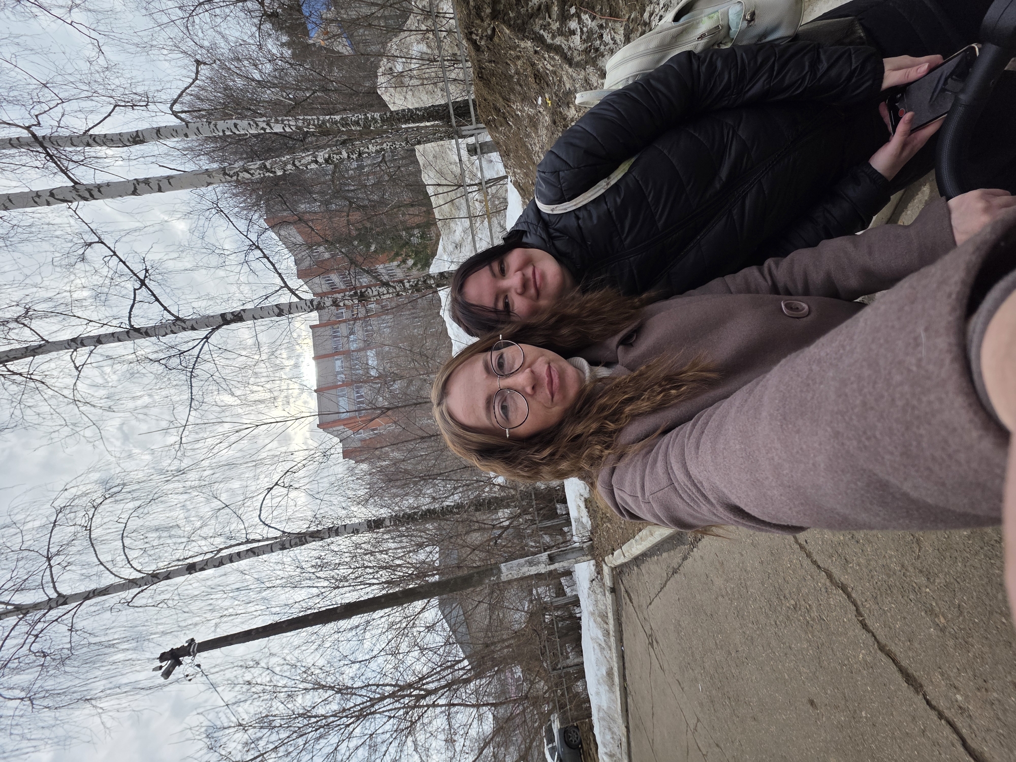 The image depicts two adult females taking a selfie outdoors in Chaikovsky, Russia, during what appears to be late winter or early spring. The woman on the left, with long brown wavy hair and round glasses, wears a light brown coat over a white turtleneck. She holds the camera, and her left hand is visible near a black stroller handle. Beside her, on the right, a second woman with dark hair smiles, dressed in a black puffer jacket. She holds a black smartphone with both hands, resting them on the same stroller handle. A light-colored backpack is visible on her right shoulder.
The ground in the immediate foreground consists of cracked asphalt to the left, transitioning to a dirt path and a snowy, sloped embankment on the right. A metal wire fence runs horizontally across the mid-ground, partially obscured by dirty snow and earth. Behind the subjects and the fence, numerous leafless deciduous trees, including several distinctive birch trees with white bark, are prominent. In the background, multiple multi-story residential buildings, characterized by their reddish-brown and gray facades, are visible through the bare tree branches. A utility pole with cross-arms stands on the far left. A dark-colored vehicle is partially visible parked on a snow-covered area further back on the left. The sky is overcast with gray clouds.