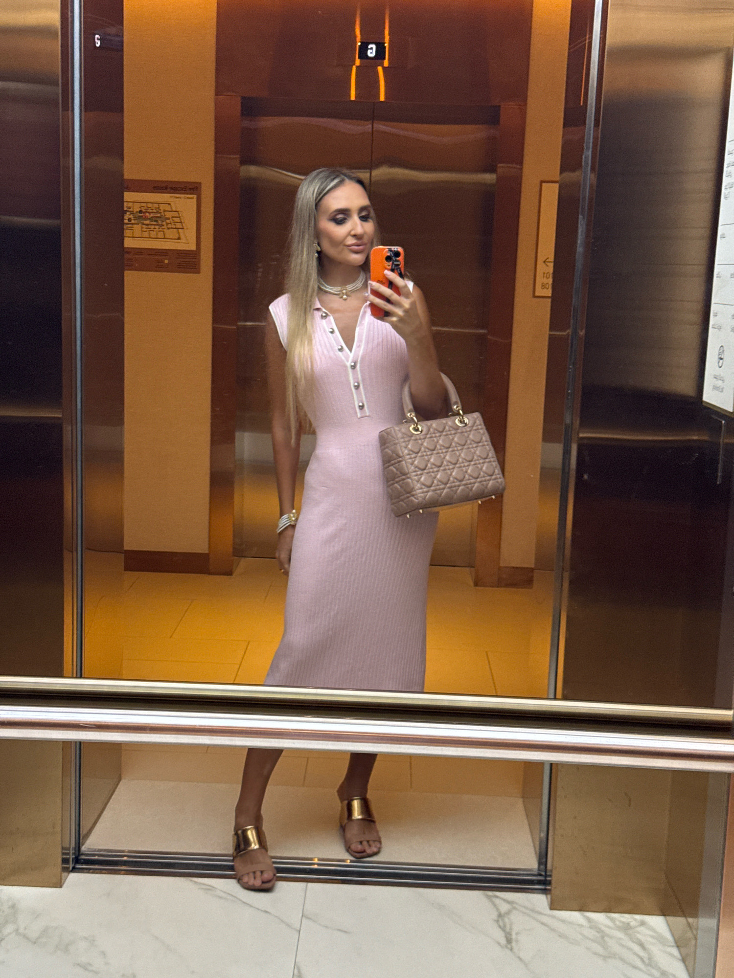 A woman stands in an elevator, taking a selfie in the reflective doors. She is wearing a light pink, ribbed, midi-length dress with a collared neckline and a button-up front. She accessorizes with a pearl necklace, a gold bracelet, and a light beige quilted handbag with gold hardware. She is also wearing gold open-toe heeled sandals. The elevator's interior is paneled with mirrored and warm-toned wood surfaces. A building directory or map is visible on one of the walls. The floor outside the elevator appears to be a patterned tile. The overall impression is one of a stylish individual in a modern urban setting, possibly in Dubai, United Arab Emirates, a city known for its contemporary architecture and luxury environments.