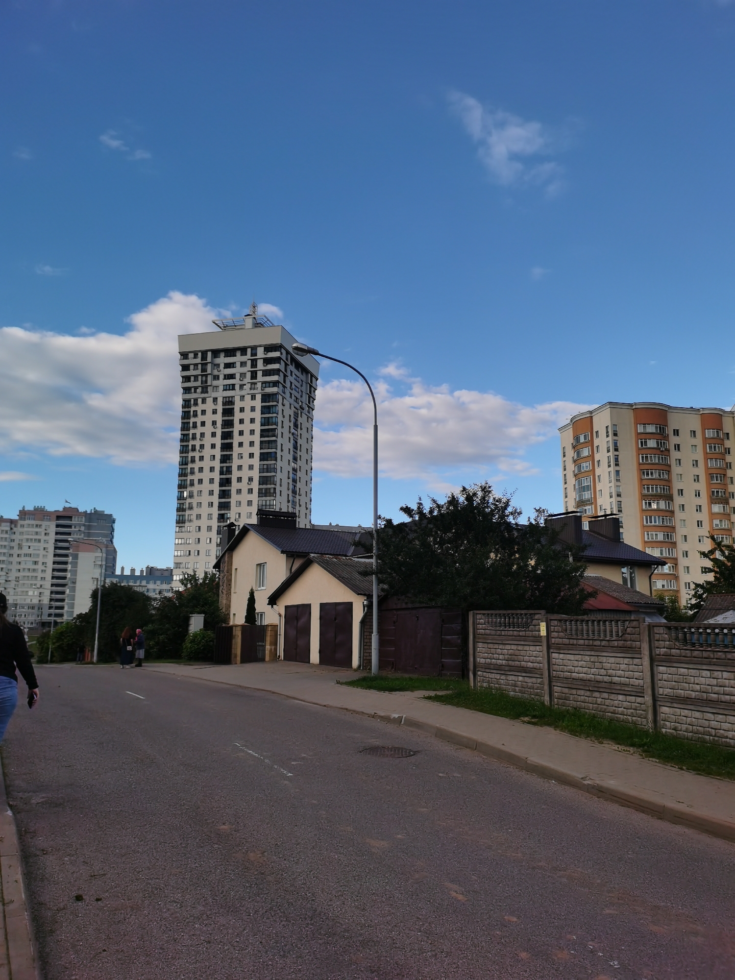 The image shows a street scene in Minsk, Belarus. The foreground features an asphalt road with a sidewalk on the right. A person is partially visible on the left, walking away from the camera. In the mid-ground, there are several buildings, including a tall, modern apartment building with a white facade and numerous windows, and a smaller, older building with a light-colored facade and brown garage doors. Behind these, other apartment buildings of various architectural styles are visible. A stone fence with patterned caps runs along the right side of the sidewalk. The sky is blue with scattered white clouds. A lamppost is visible on the right side of the road, with its curved arm extending over the sidewalk. The overall impression is of a residential or mixed-use urban environment.