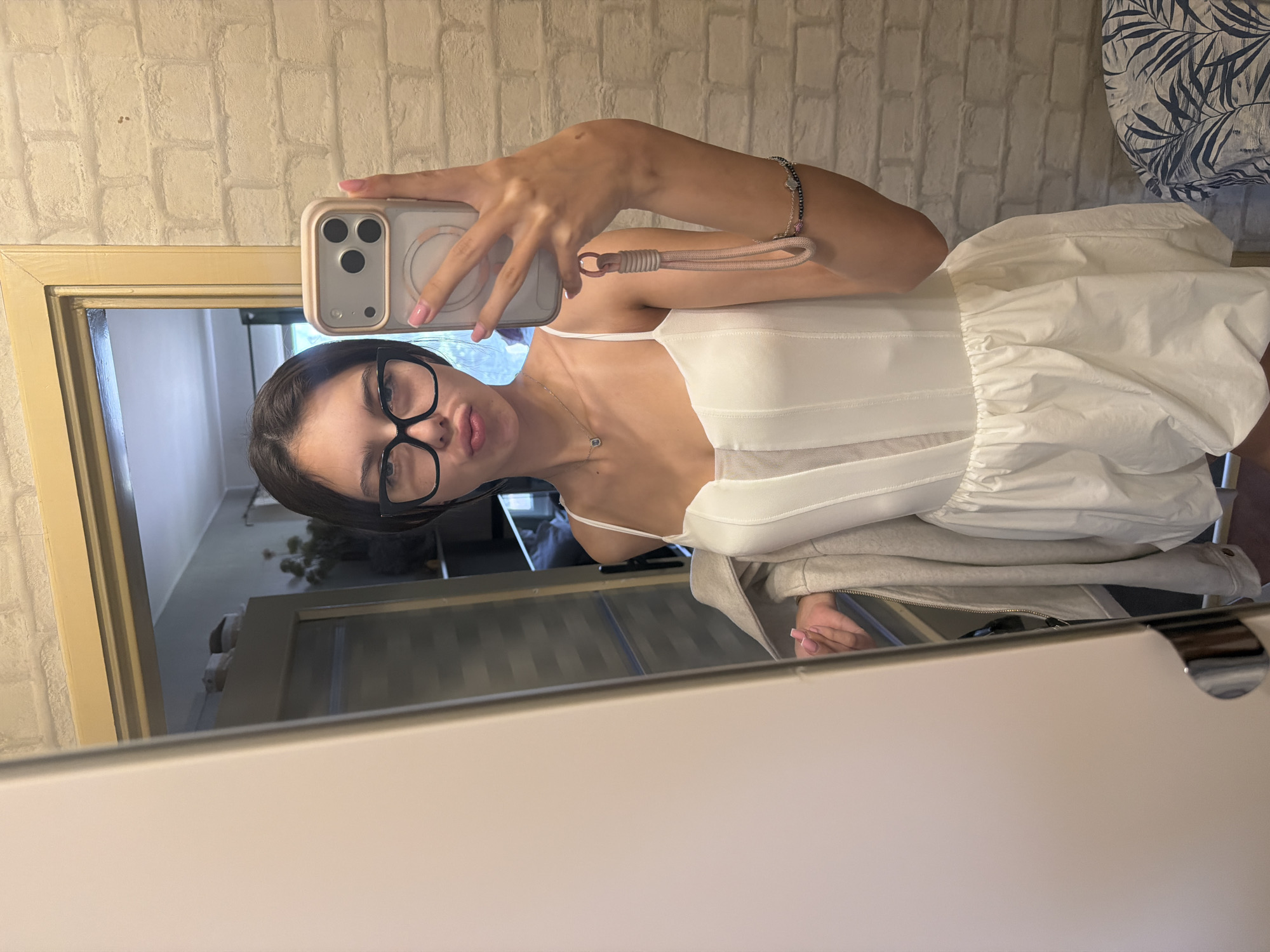 This is a vertically oriented, medium shot selfie taken in a room, possibly a bedroom. The subject is a young woman with dark hair and glasses, wearing a white camisole dress and a light grey cardigan. She is holding up a smartphone to take the picture, with her left hand visible. A bracelet adorns her left wrist. The background consists of a brick-patterned wallpaper and a mirror that reflects the room. The setting appears to be indoors, with a door and a piece of furniture visible in the reflection. The lighting suggests it could be daytime, but the exact time is unclear. The woman's expression is neutral. There is no visible text in the image.