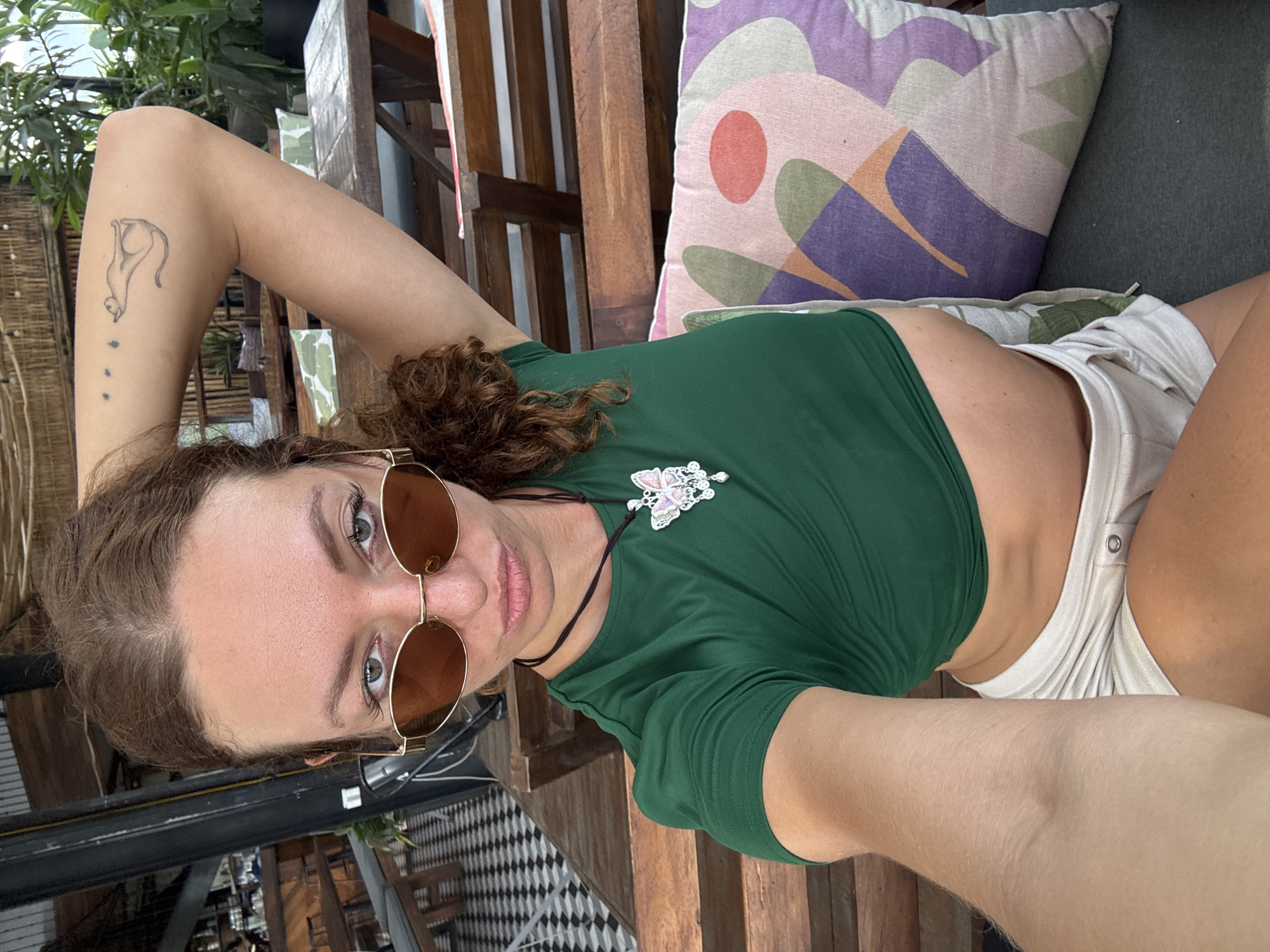 This well-lit, sideway-oriented selfie captures a young woman relaxing in an outdoor or semi-open setting during daytime. She lies diagonally across the frame, looking directly at the camera with a calm expression.
The woman has brown, curly hair and fair skin. Her light-colored eyes are visible above gold-rimmed, round sunglasses that rest on her nose. She is dressed in a dark green, short-sleeved crop top, exposing her midriff, and light-colored shorts. A stylized tattoo of a cat or feline figure, with three small dots beneath it, is visible on her left upper arm. She wears a necklace with a pink and white butterfly pendant.
She appears to be reclining on wooden lounge furniture, with wooden slats visible around her. A patterned pillow featuring abstract shapes in purple, pink, and green is positioned behind her head, alongside another plain dark green cushion. The background shows green foliage, indicating an outdoor environment, and a patterned black and white floor is partially visible. The overall scene suggests a relaxed atmosphere in a cafe, restaurant, or lounge area in Hà Giang 1 Ward, Vietnam. No visible text is present in the image.